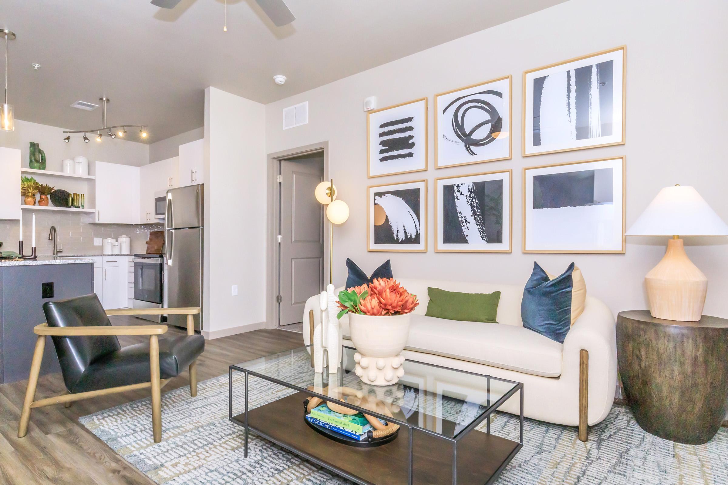 Modern living room featuring a cozy white sofa with decorative pillows, a stylish black armchair, and a coffee table with a decorative vase. The walls are adorned with framed abstract art, and there is a kitchen area visible in the background with stainless steel appliances. A warm lamp adds ambiance to the space.