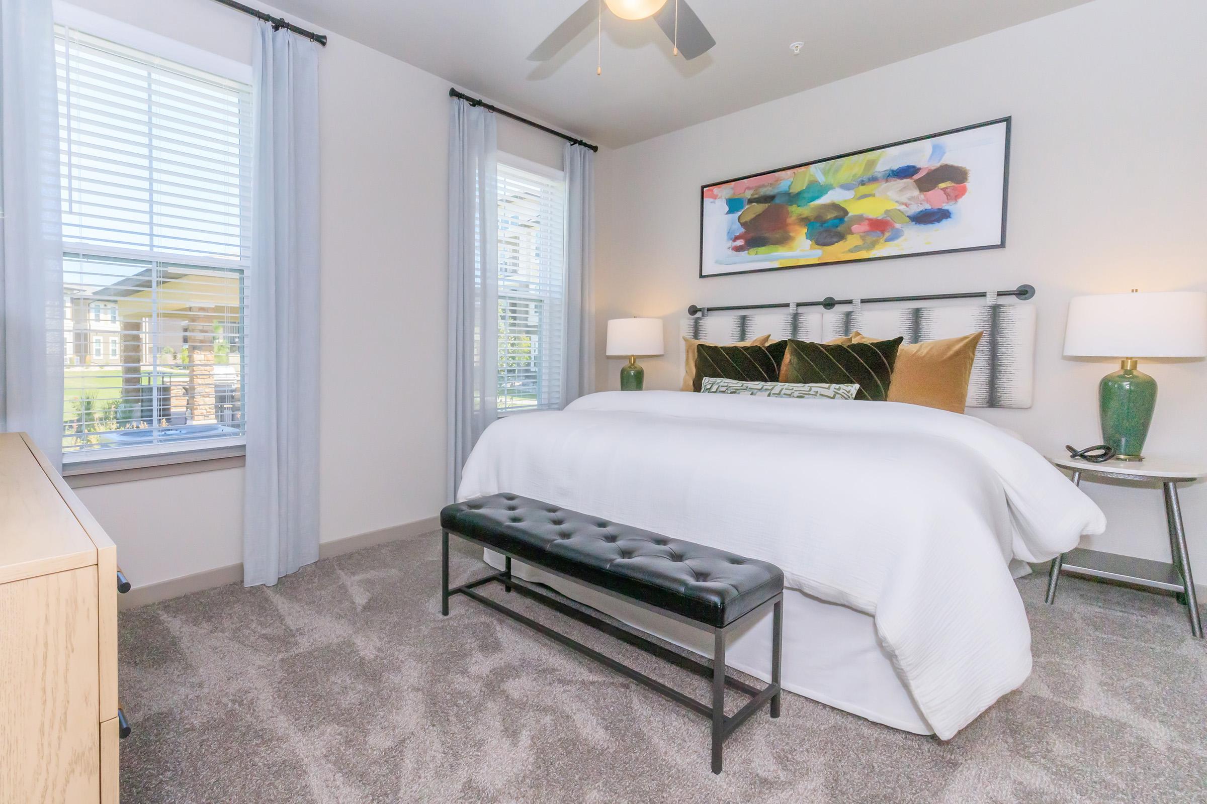 A cozy bedroom featuring a large bed with white linens, accented by colorful artwork above the headboard. Two bedside lamps provide soft lighting, and a bench at the foot of the bed adds functionality. Natural light streams in through two windows, illuminating the plush carpet.