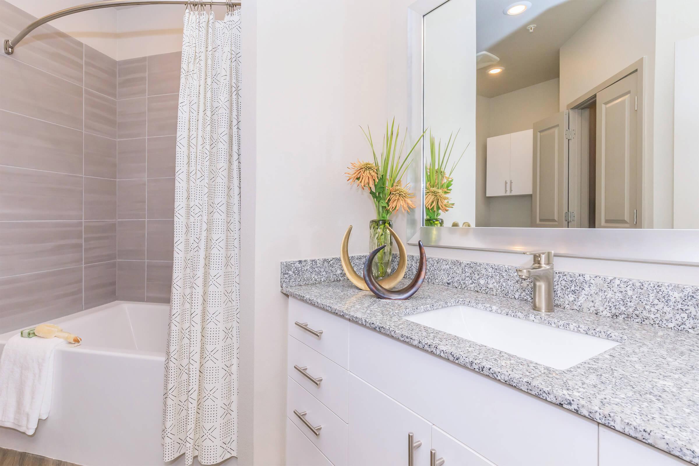 A modern bathroom featuring a bathtub with a shower curtain, a granite countertop with a sink, and decorative elements including a vase with flowers and unique sculpture pieces. Neutral-colored walls and a large mirror enhance the spacious feel.