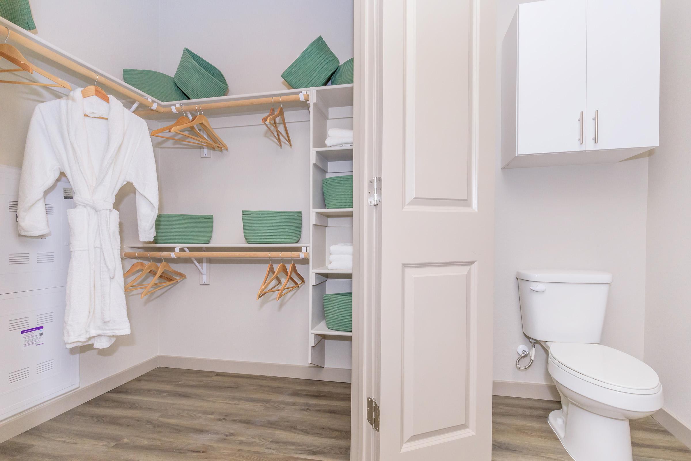 A tidy closet featuring white bathrobes hanging on wooden hangers, green woven storage baskets, neatly stacked towels, and a white toilet. The walls are painted light colors, and the flooring is a warm wood style.