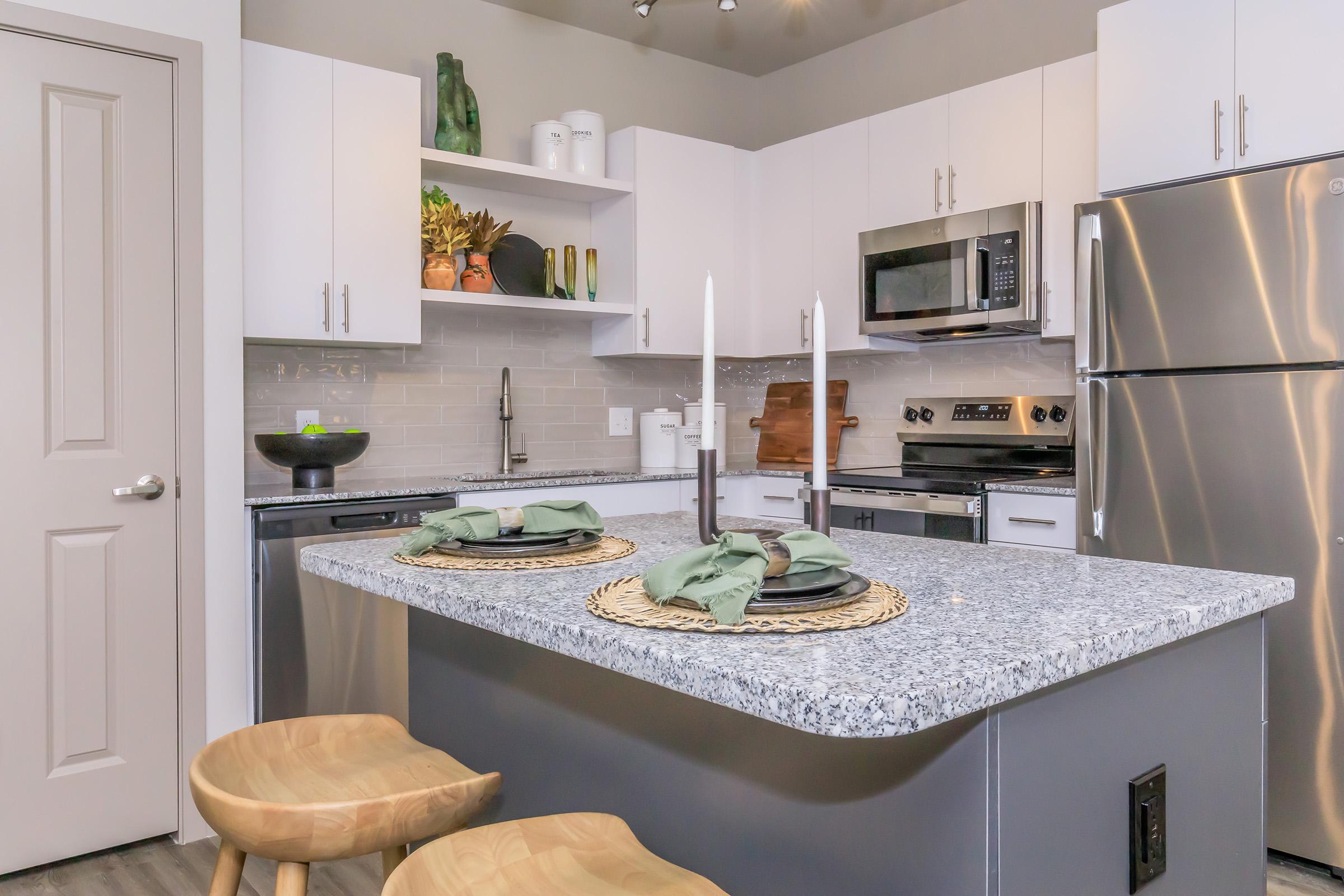 Modern kitchen featuring a gray granite countertop with two wooden stools, decorative place settings, white cabinets, stainless steel appliances, and open shelving with plants and kitchenware. Soft lighting accentuates the space, creating a warm and inviting atmosphere.