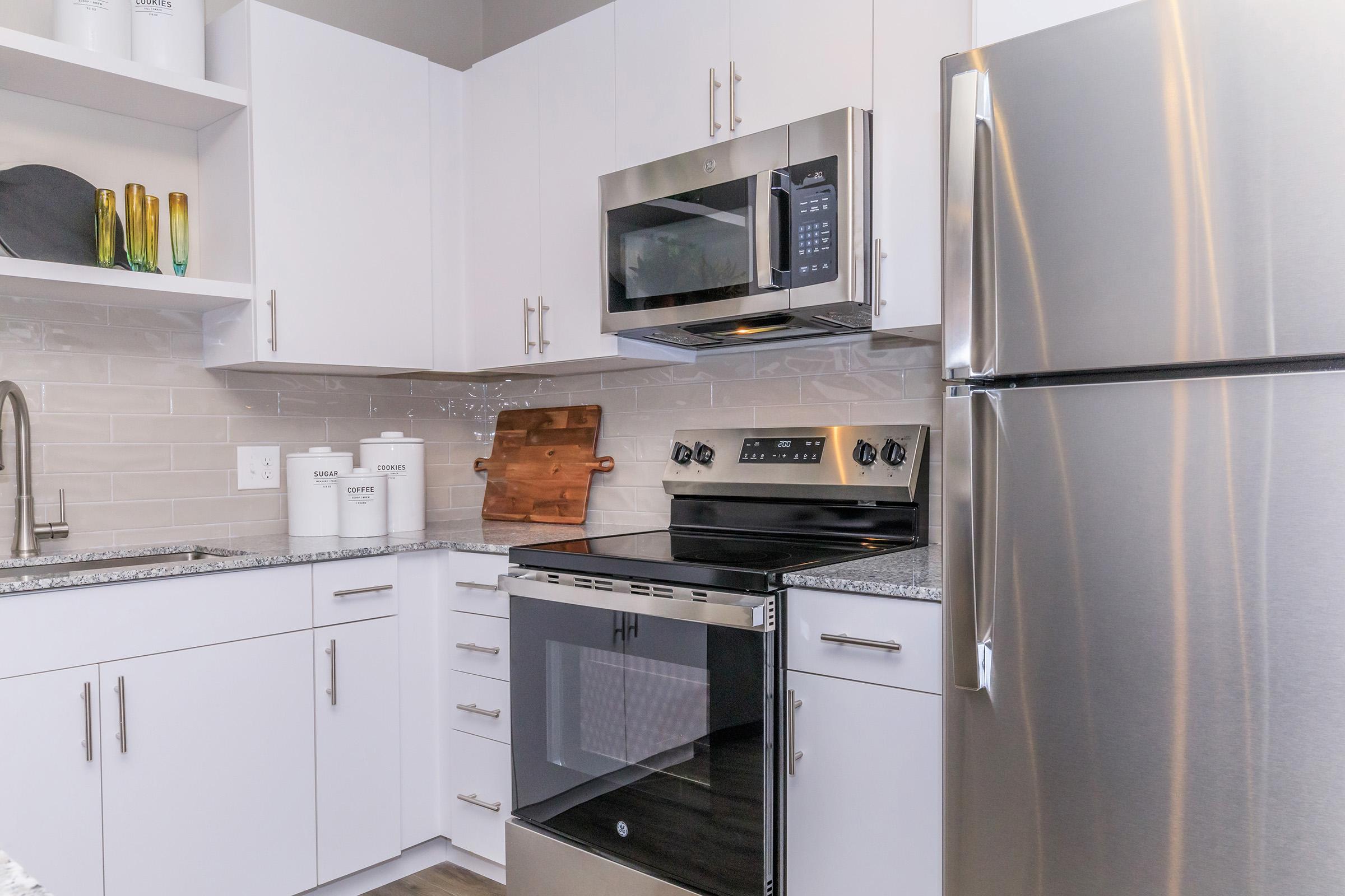 Modern kitchen featuring white cabinetry, stainless steel appliances including a microwave and oven, a sleek refrigerator, and a granite countertop. There are decorative items on the shelves and a wooden cutting board on the counter, creating a clean and functional cooking space.