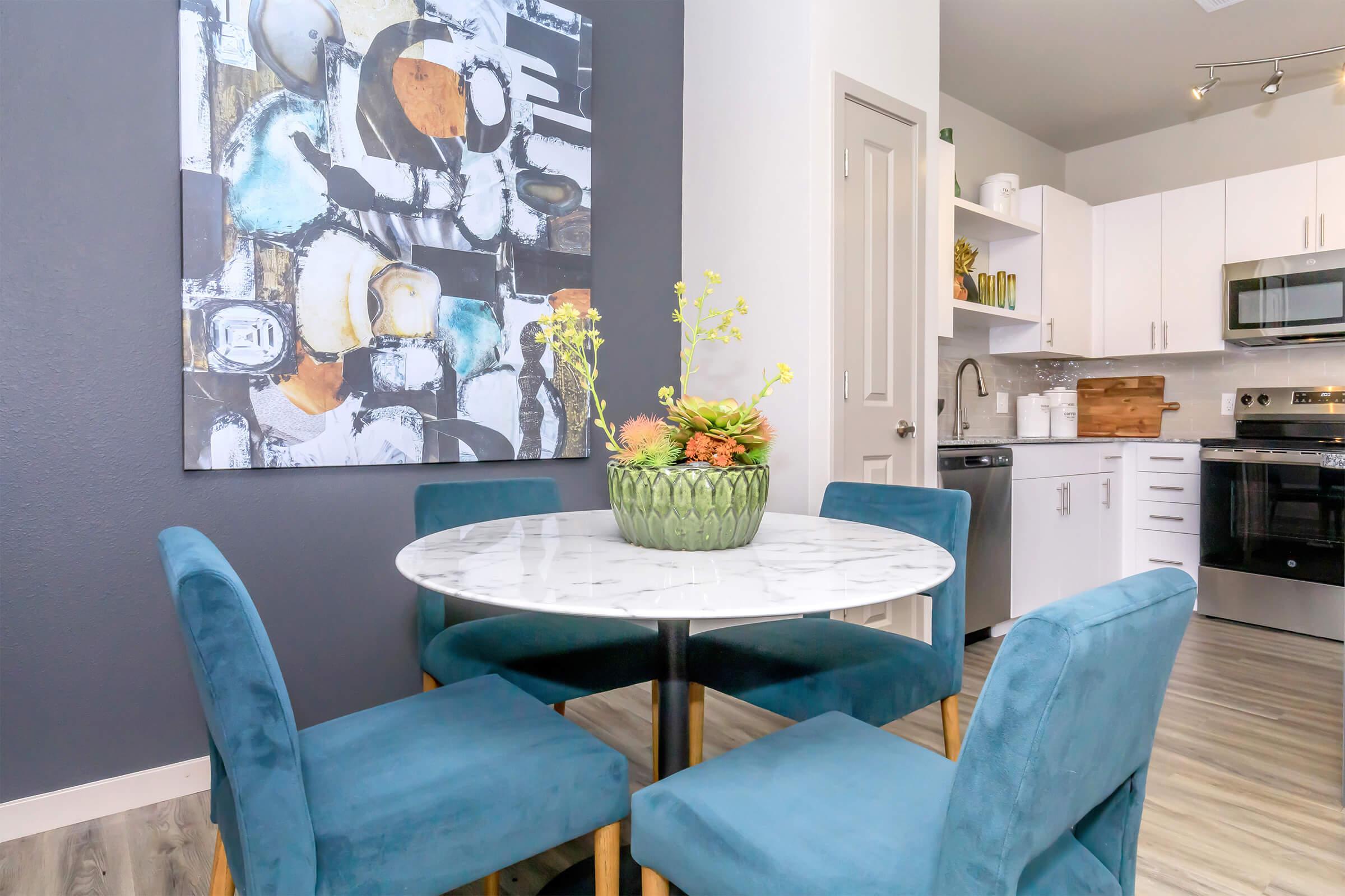 A stylish dining area featuring a round marble table surrounded by four teal chairs. A decorative green bowl with colorful flowers and plants sits at the center of the table. The wall behind is adorned with a modern abstract painting, and a light-filled kitchen area is visible in the background.