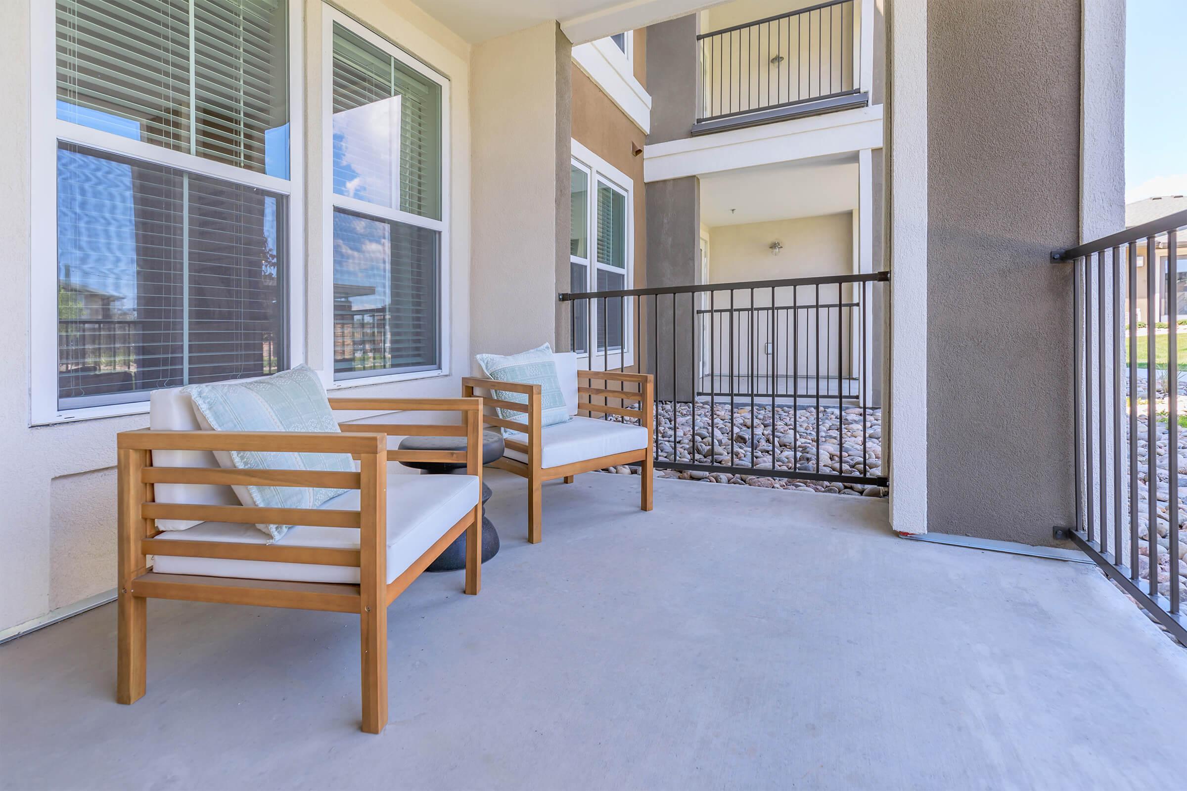 A cozy outdoor patio area featuring two modern wooden chairs with white cushions, positioned on a concrete floor. The backdrop includes large windows of an apartment building, allowing natural light to enhance the space. A metal railing surrounds the patio, with a neat gravel area visible below.