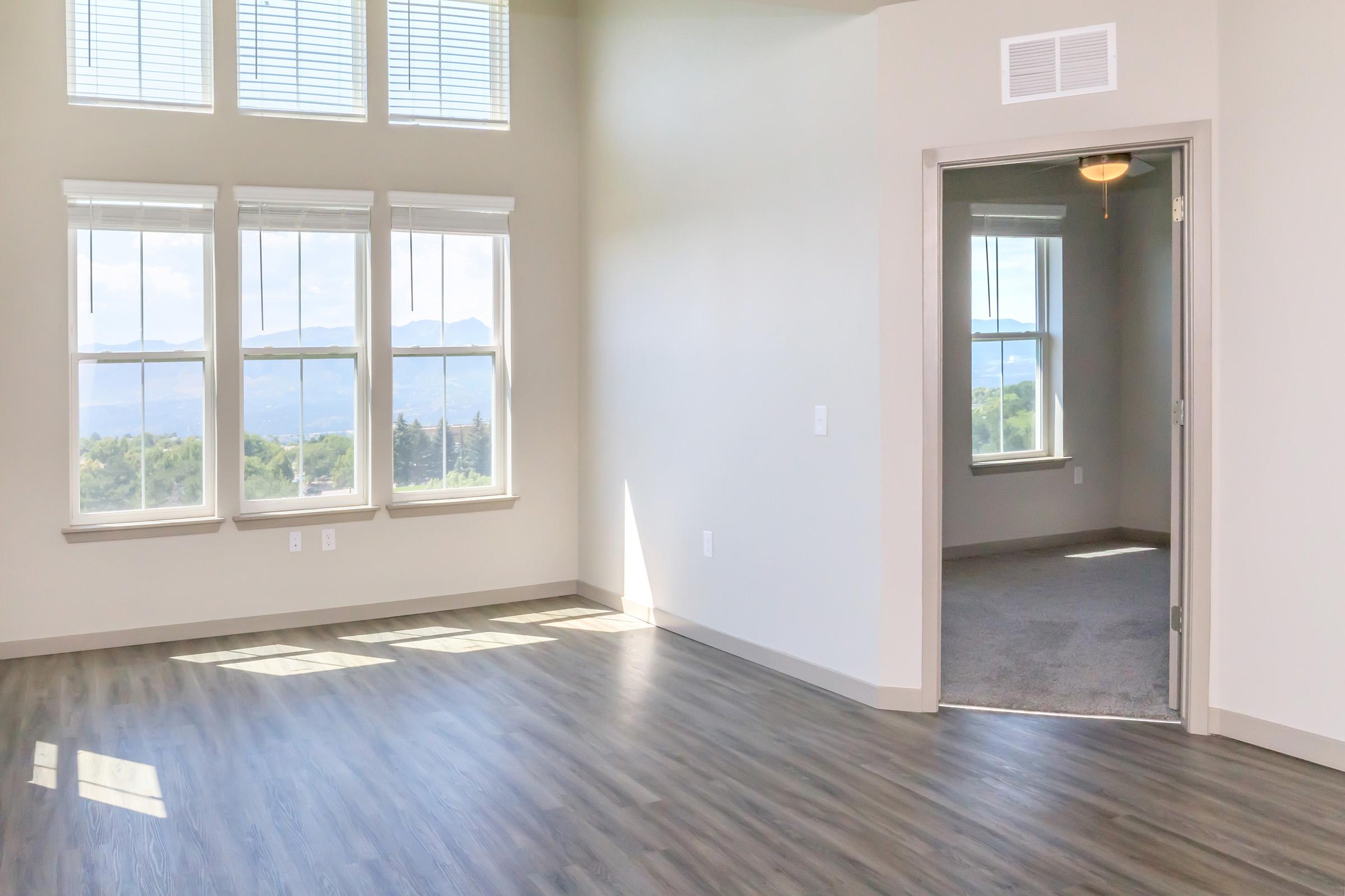 Spacious interior of a modern room featuring large windows with natural light, hardwood flooring, and a doorway leading to an adjoining room. The walls are painted in light colors, creating a bright and airy atmosphere. Scenic views of mountains are visible through the windows.