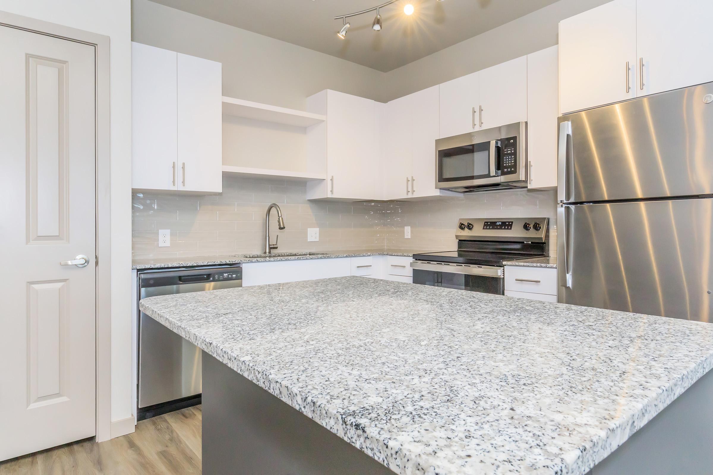 Modern kitchen featuring white cabinets, stainless steel appliances, and a large granite island. The kitchen is well-lit with overhead lighting and a neutral color palette, creating a spacious and inviting atmosphere.