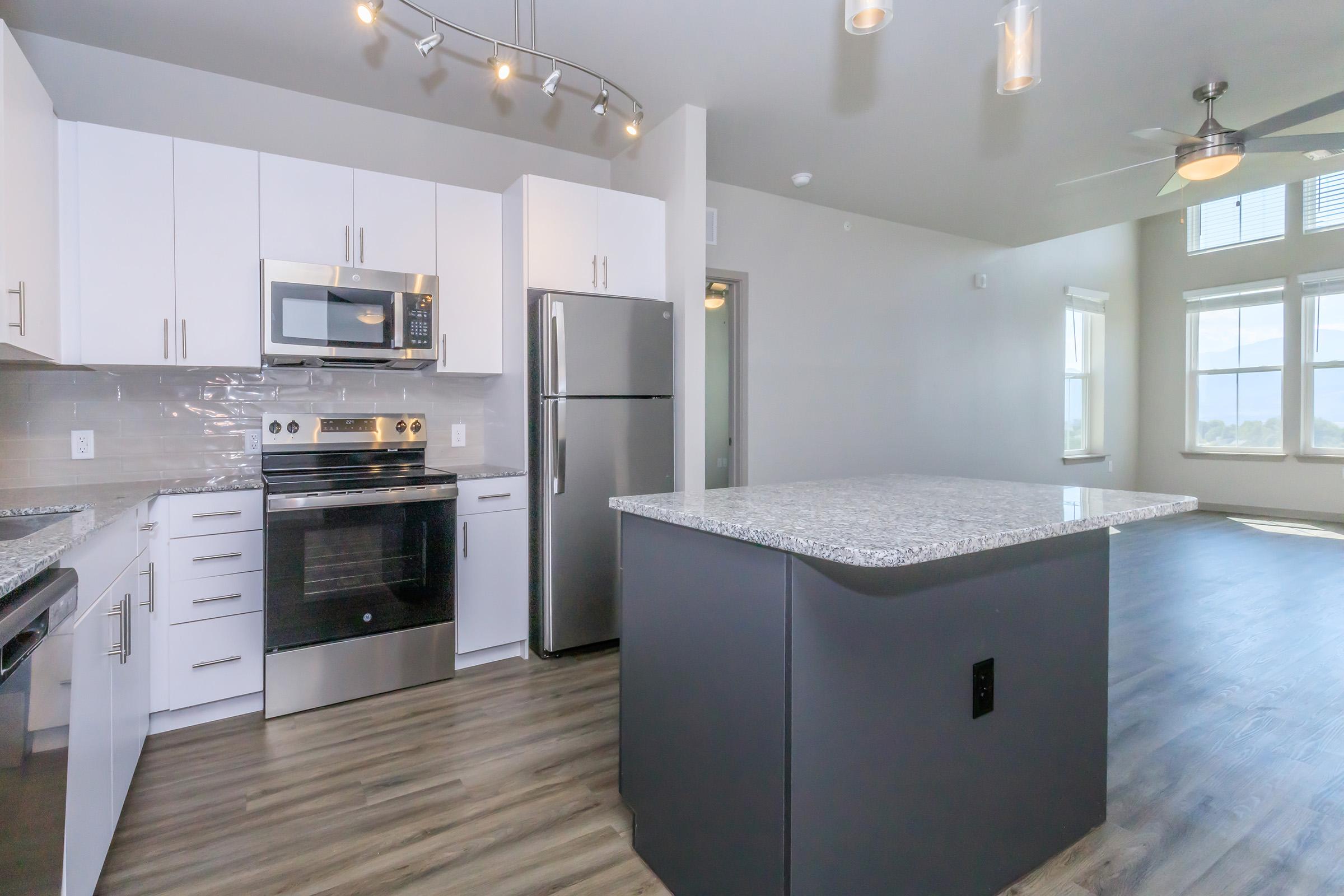 Modern kitchen featuring white cabinets, stainless steel appliances including a microwave and refrigerator, a large granite island, and wood-like flooring. Bright natural light streams through large windows, creating an open and inviting space.