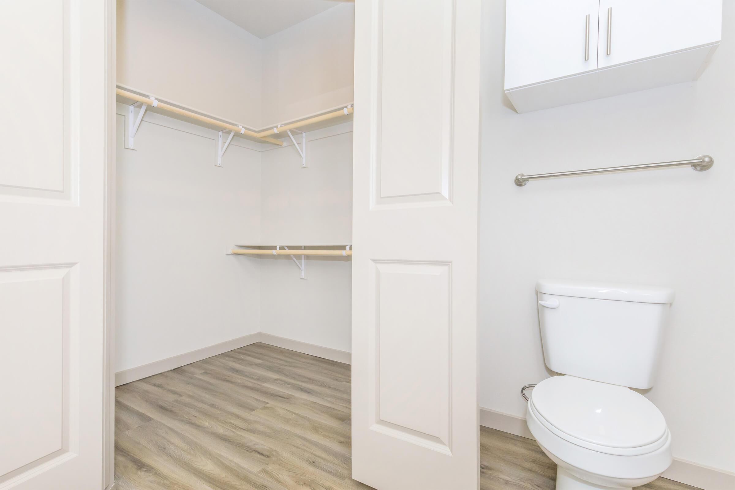 Interior of a closet space featuring two shelves mounted on the wall, a white toilet positioned nearby, and light-colored flooring. The walls are painted in a neutral shade, enhancing the open and airy feel of the area.