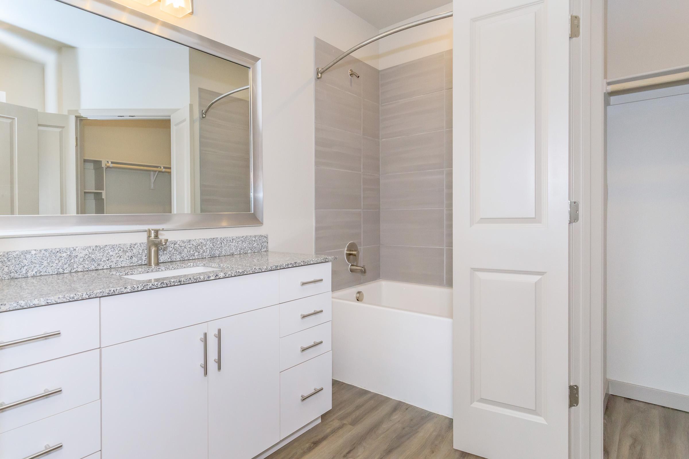 Bright and modern bathroom featuring a white granite countertop with sleek cabinetry, a large mirror, a bathtub with a showerhead, and light-colored tile walls. The space has light wood flooring and is well-lit by natural light. A closet is visible in the background.