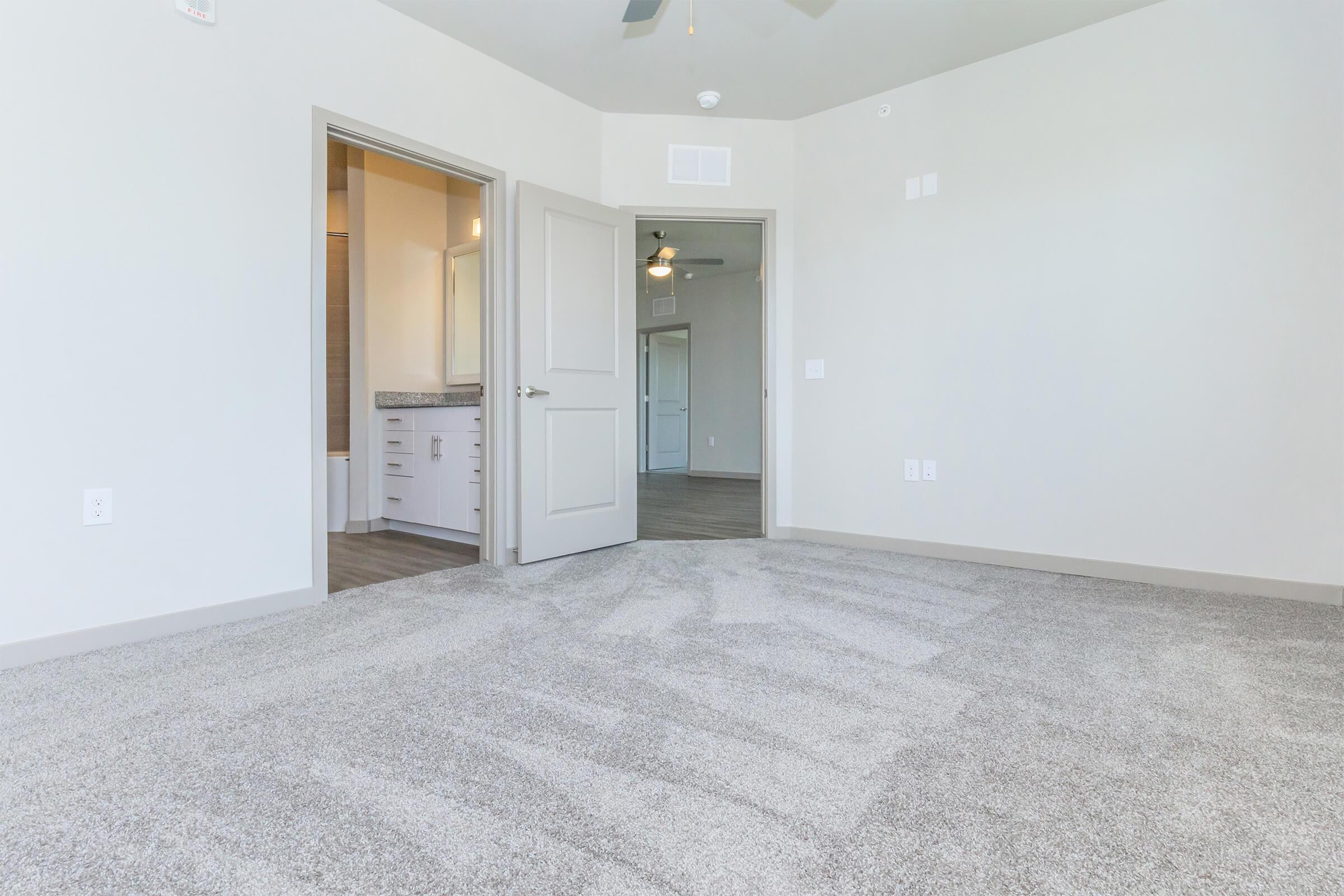 A spacious, empty room with light gray carpet and painted walls. A doorway leads to a bathroom area, and there's a ceiling fan overhead. Soft natural light fills the room, creating an airy feel. The design is modern and minimalist, ideal for various uses or decoration.