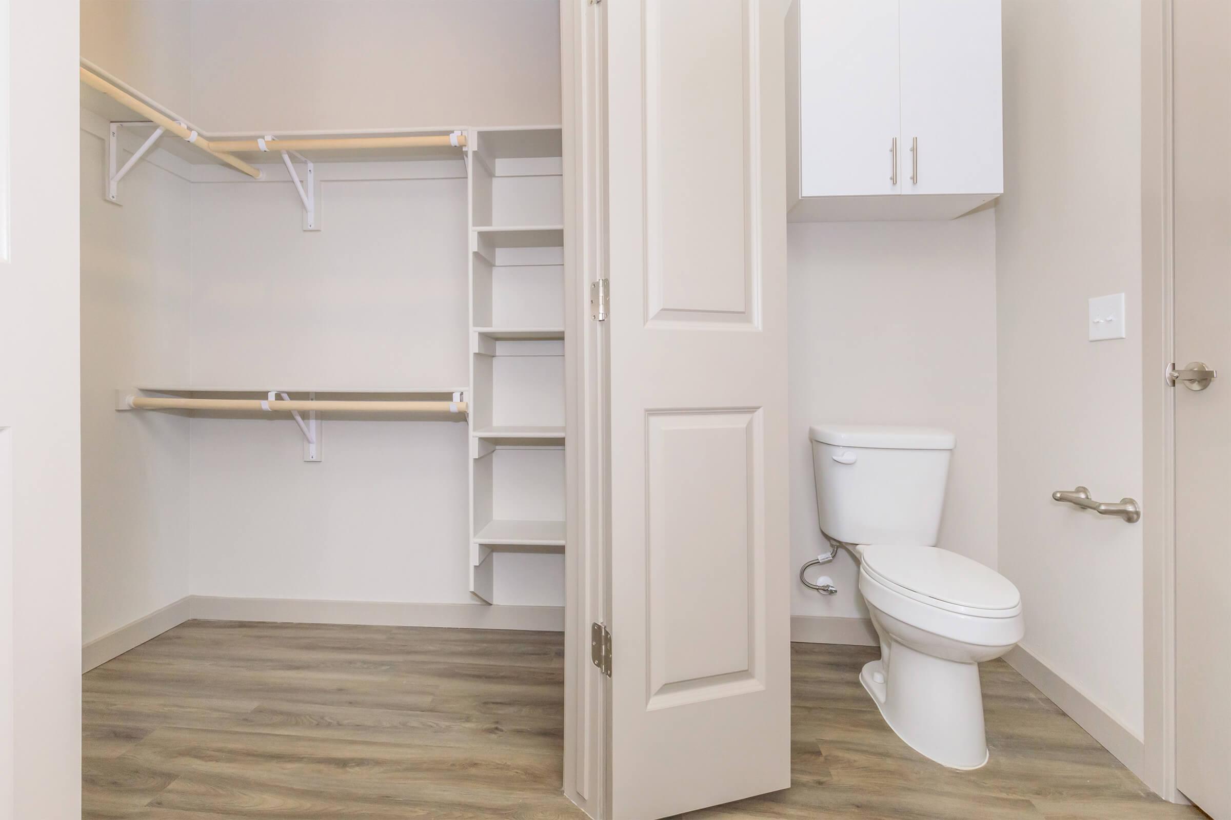 A tidy, modern small space featuring a closet with open shelving and hanging rods on the left, a toilet on the right, and light-colored walls. The flooring is a warm wood tone, and there is a light cabinet above the closet shelves.