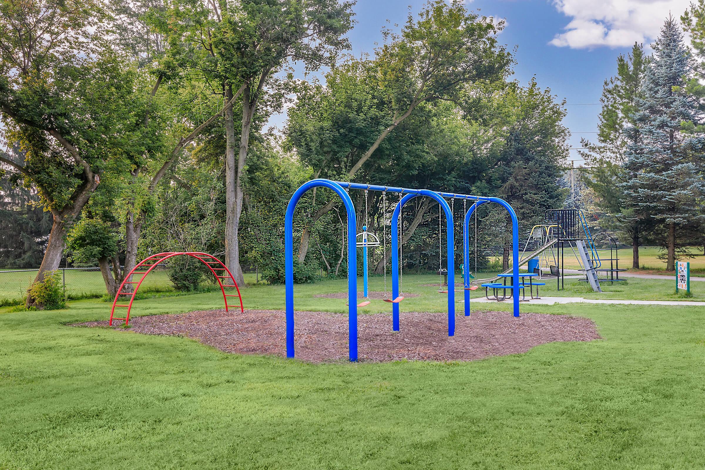 A colorful playground featuring blue swings, a red slide, and a climbing structure surrounded by lush green grass and trees under a clear blue sky. The area is well-maintained, ideal for children to play and explore.