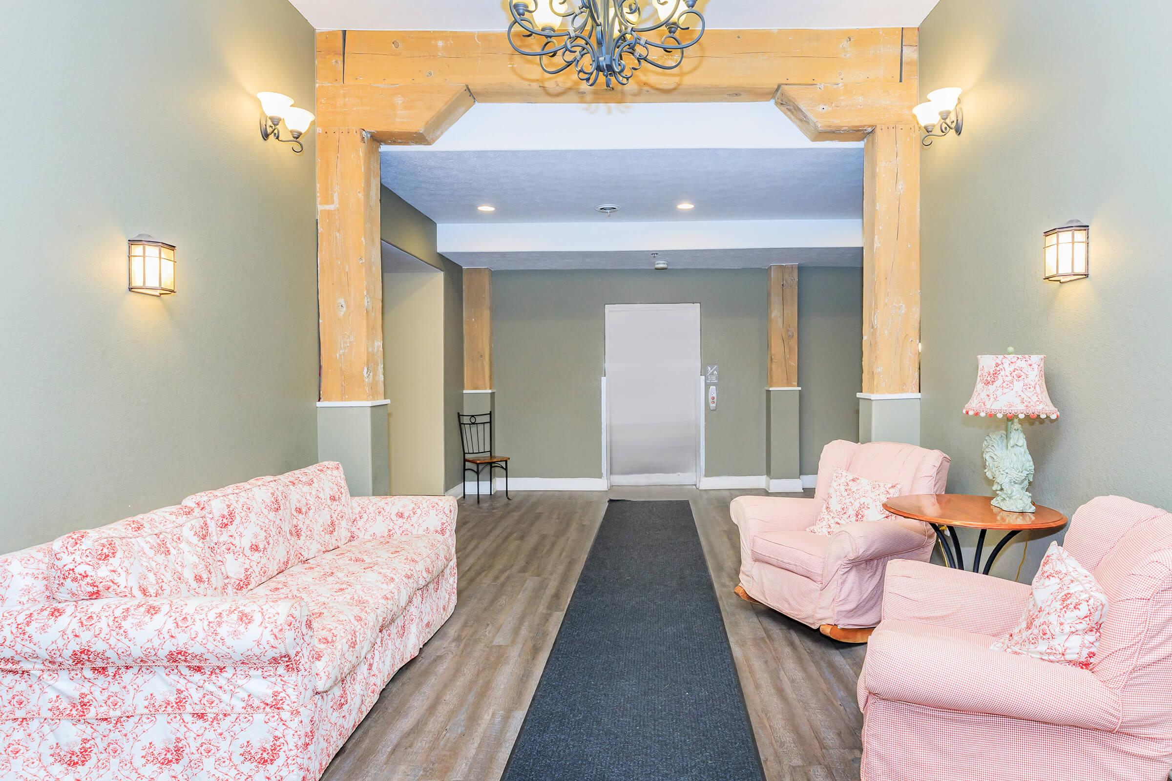 A well-lit hallway featuring two floral-patterned sofas, one pink armchair, and a small wooden table with a lamp. The walls are painted green, adorned with wall sconces, and wooden beams accent the entrance area. A carpeted runner leads towards an elevator at the end of the hallway.
