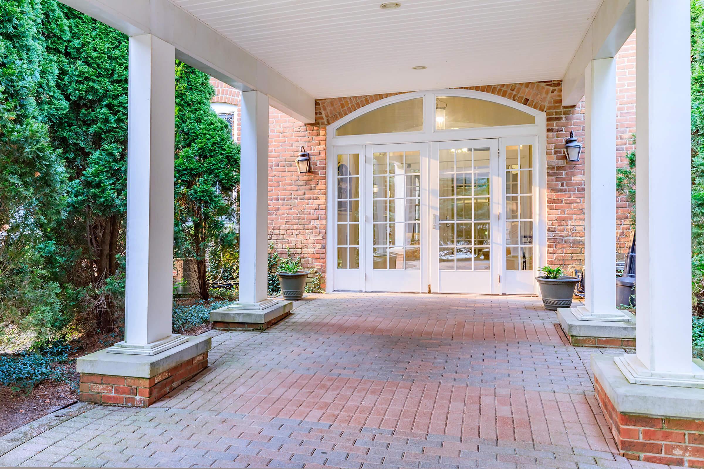 A welcoming entrance with a paved walkway leading to a set of double glass doors, surrounded by brick walls and lush green shrubbery. Two decorative lanterns are mounted on the columns, and potted plants are placed on either side of the doors, creating an inviting atmosphere.