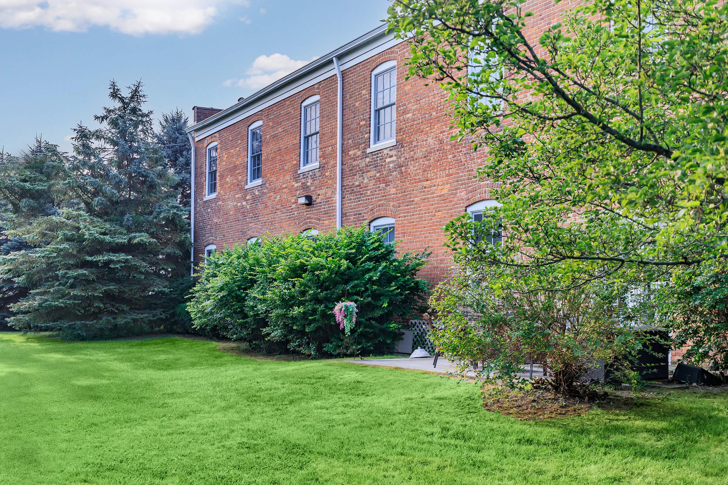 A brick building with large windows stands next to a lush green lawn. Surrounding the building are trees and shrubs, creating a serene outdoor space. The sky is partially cloudy, adding to the peaceful atmosphere of the scene.