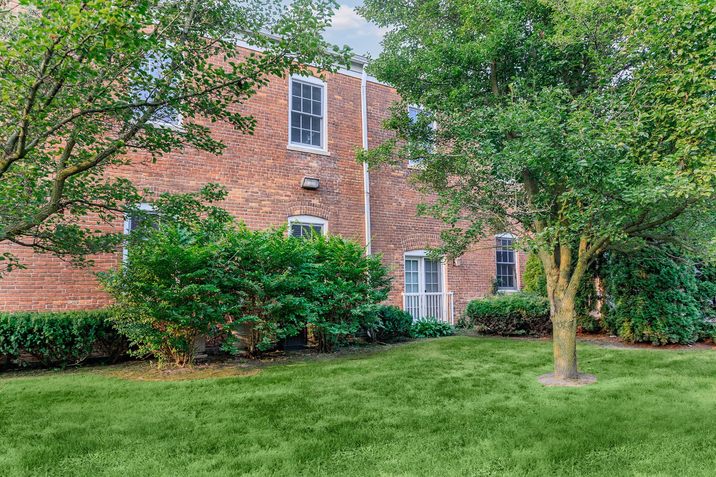 A brick building surrounded by lush greenery, including shrubs and trees, with a well-maintained lawn in front. The scene conveys a serene outdoor setting, with windows visible on the building's facade.