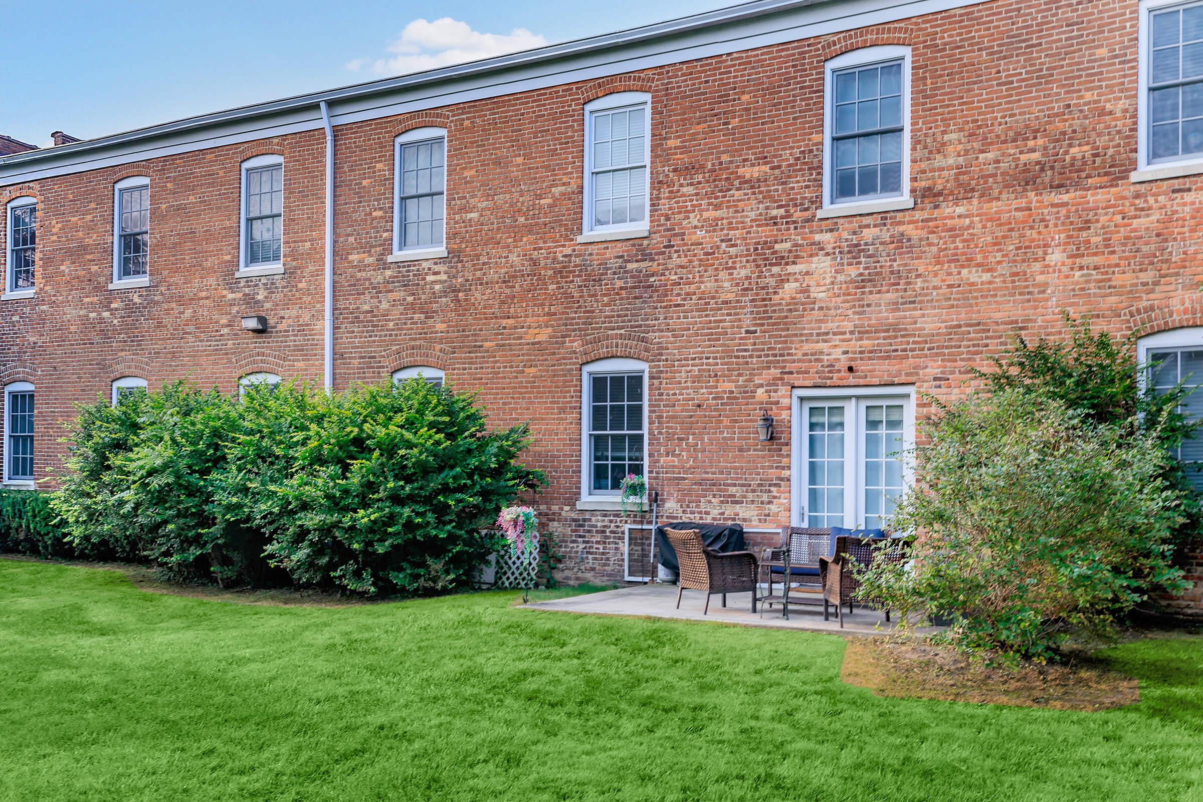 A brick building with multiple windows, featuring a small patio area with outdoor furniture. Green hedges and grass surround the patio, creating a welcoming outdoor space. The setting is bright and well-maintained, suggesting a pleasant residential environment.