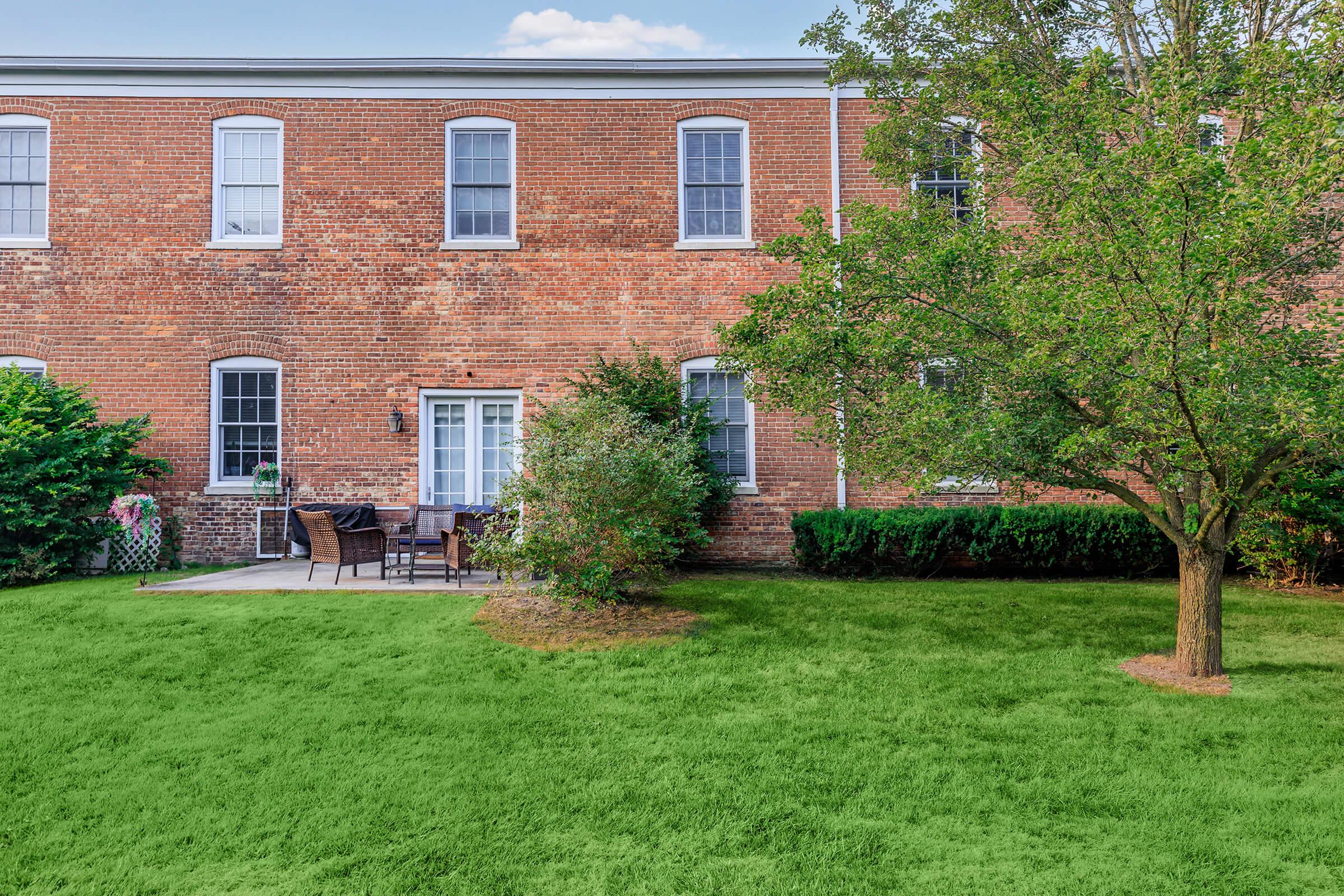 A brick building with multiple windows, featuring a small patio area with outdoor seating. Surrounding the patio is a lush green lawn and a small tree. The scene is peaceful and well-maintained, suggesting a residential setting.