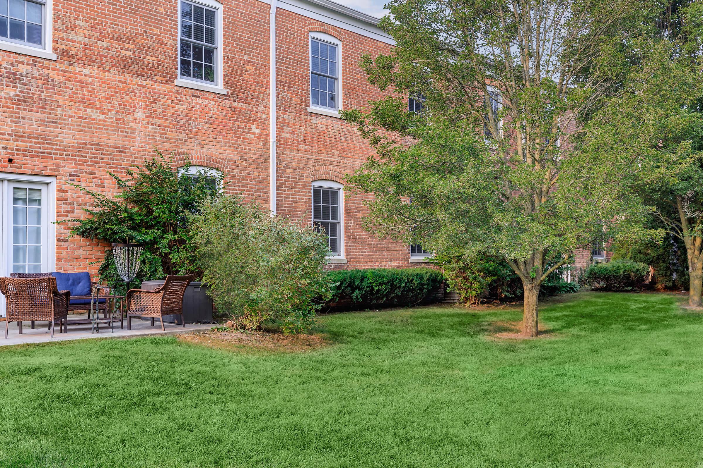 A well-maintained lawn in front of a brick building, featuring a couple of trees and shrubs. There are patio chairs and a small table set up on a concrete patio, creating an inviting outdoor space. The surroundings are lush and green, enhancing the tranquility of the setting.