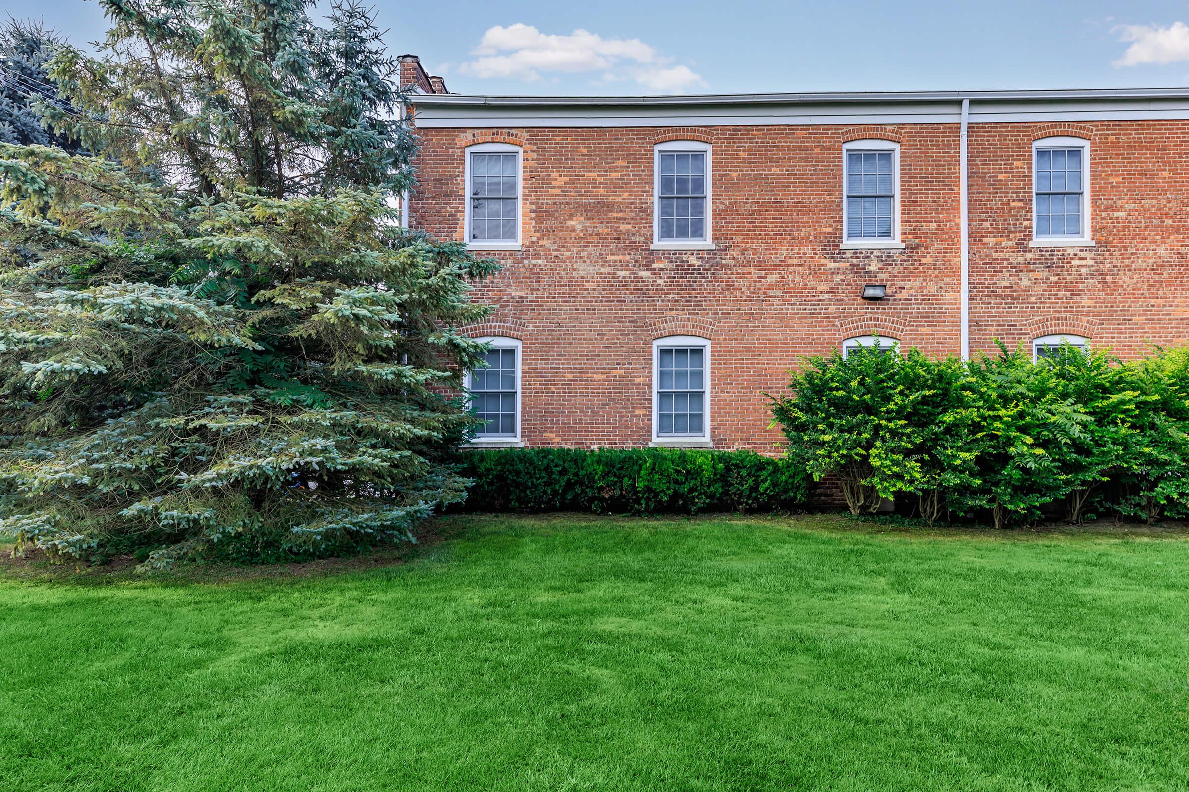 A brick building with multiple windows, surrounded by well-maintained shrubs and trees. The grassy lawn in front is lush and evenly trimmed, creating a pleasant outdoor space. Cloudy blue sky is partially visible above the structure.