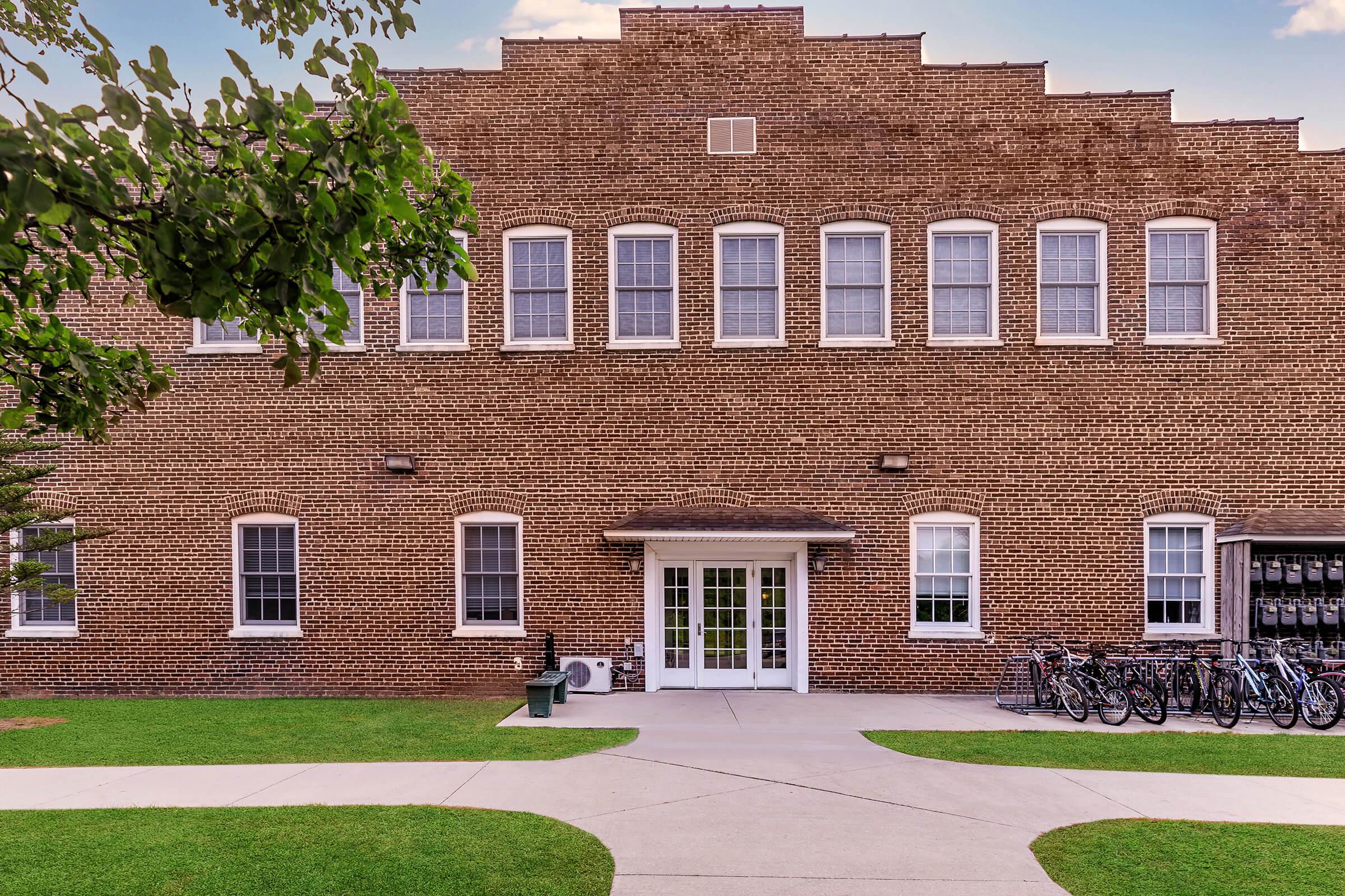 A brick building featuring multiple windows, a central entrance with double doors, and a grassy area in front. Bicycles are parked in racks to the right, while a small bench is visible in the foreground. The building has a simple, symmetrical architectural design with a stepped roofline.