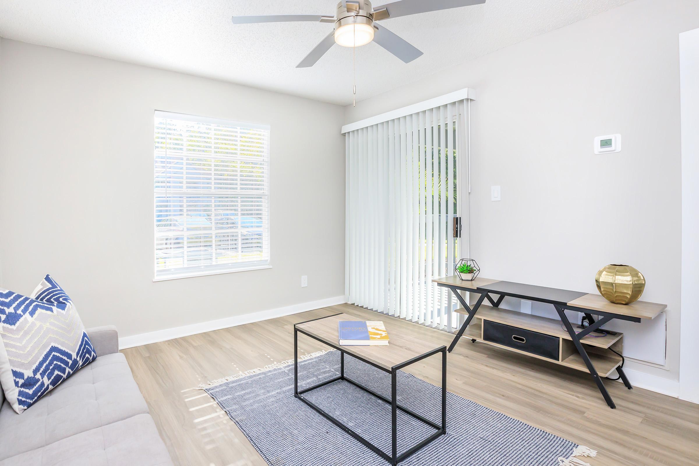 A modern, light-colored living room featuring a gray sofa with a blue patterned throw pillow, a black coffee table with a decorative book, and a minimalist TV stand. There are vertical blinds on the window, a ceiling fan above, and a cozy atmosphere created by neutral tones and natural light.
