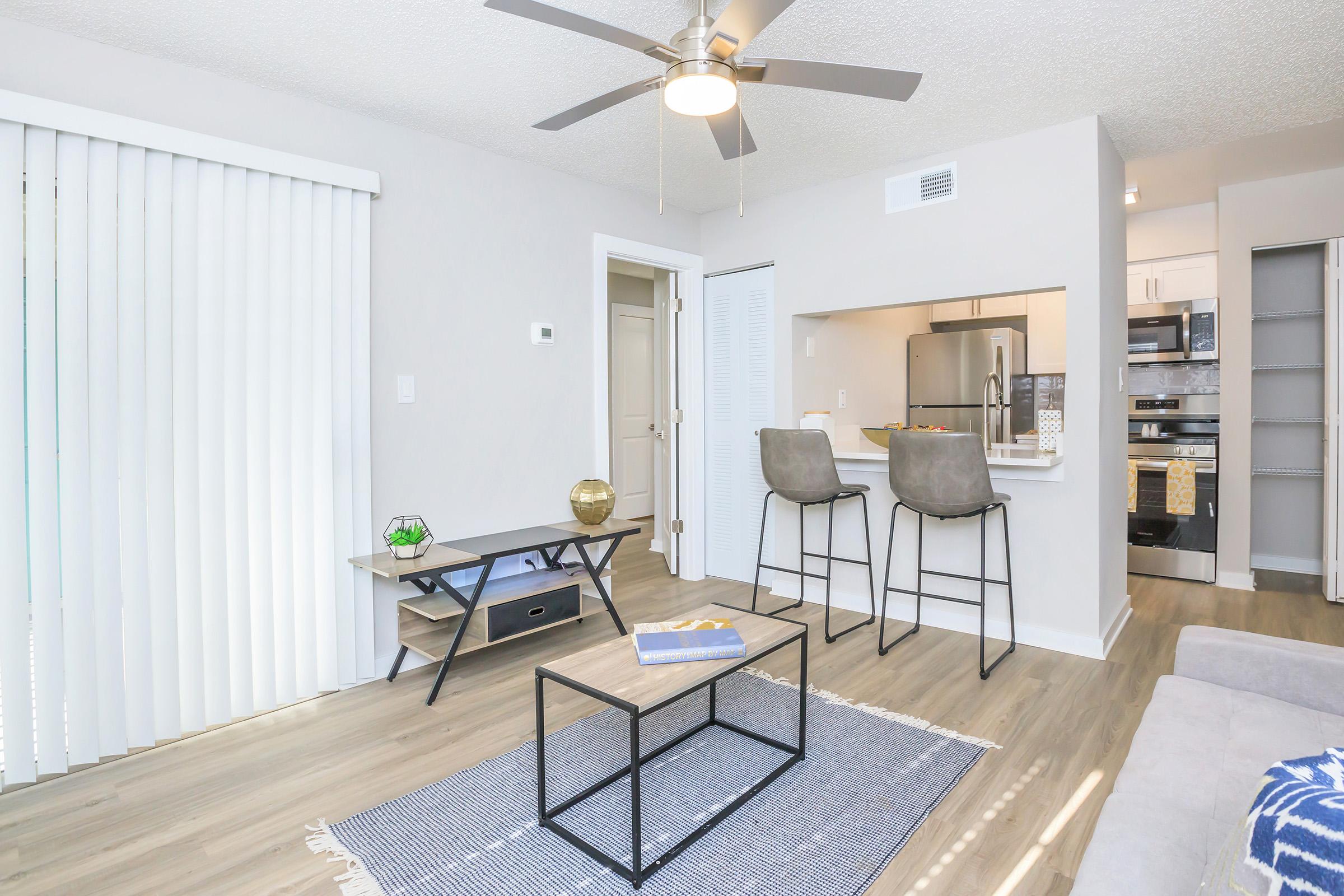 A modern living space featuring light-colored walls, a ceiling fan, and a beige sofa. There’s a small coffee table on a rug, with a TV stand and decorative items. The kitchen is partially visible with stainless steel appliances, and there are two bar stools at a counter. 