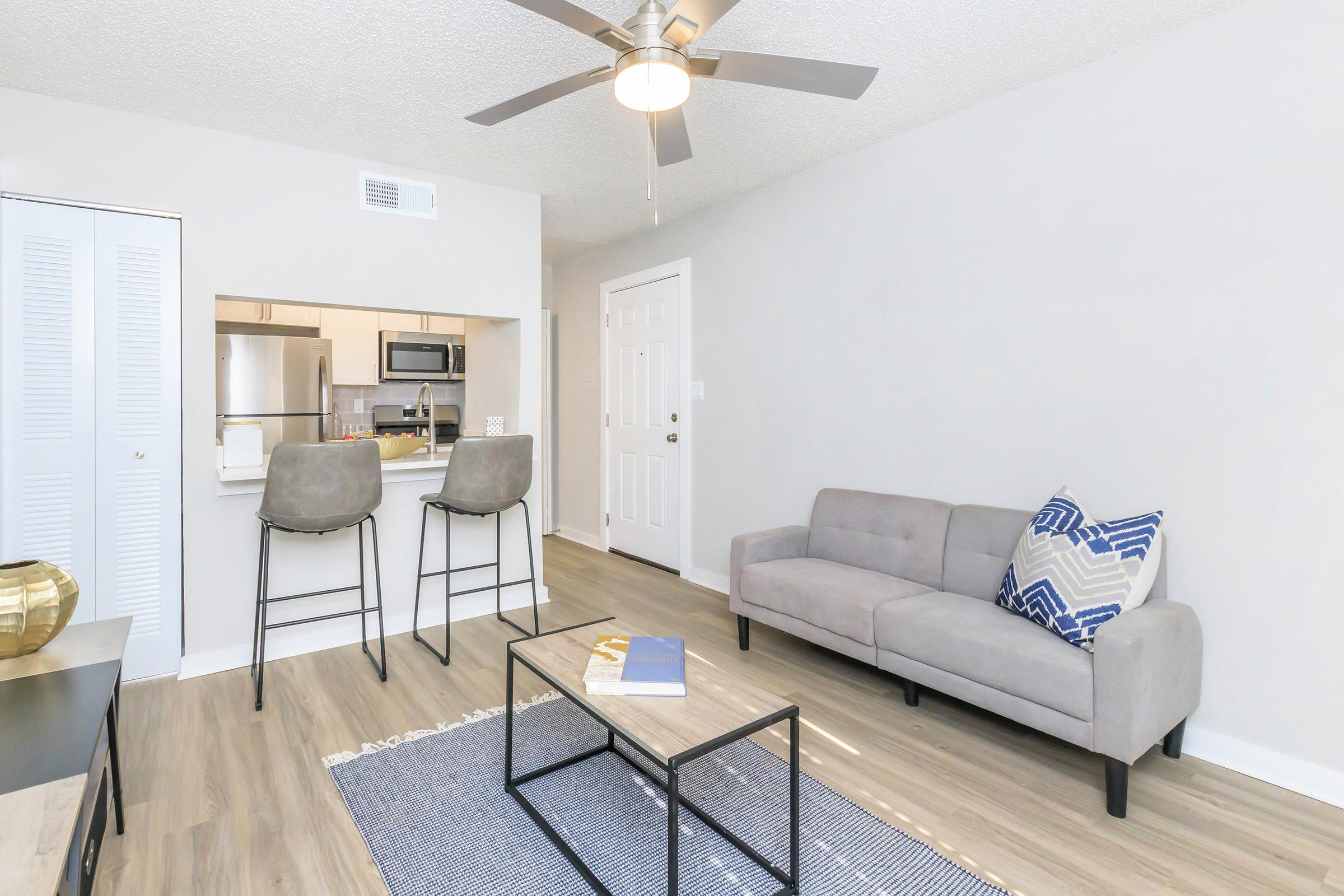 Modern living room featuring a gray sofa with a decorative pillow, a coffee table on a textured rug, and two bar stools at a kitchen counter in the background. A ceiling fan is visible, and natural light fills the space through a nearby door. The walls are painted in light colors, creating a bright and inviting atmosphere.