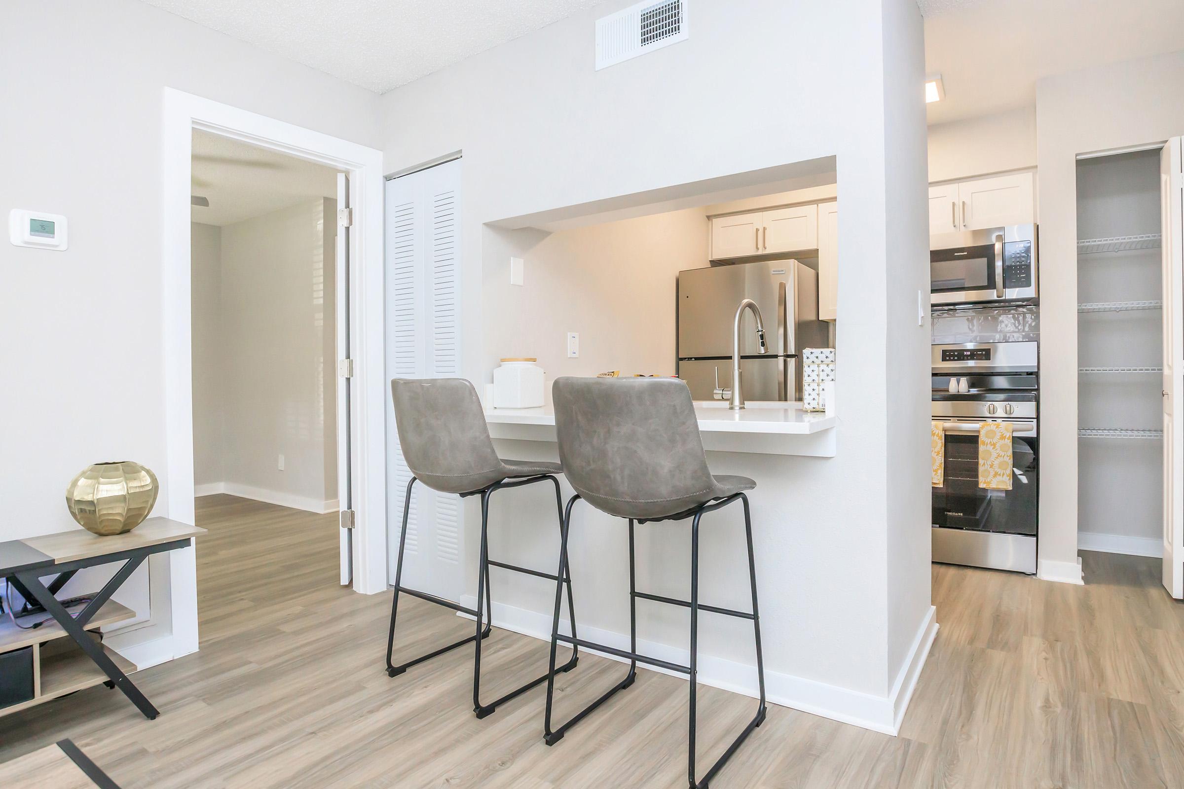 Modern kitchen and dining area featuring a breakfast bar with two gray stools, stainless steel appliances, and light-colored cabinetry. The space has neutral-colored walls and laminate flooring, with a glimpse of a living area in the background. Natural light enhances the inviting atmosphere.