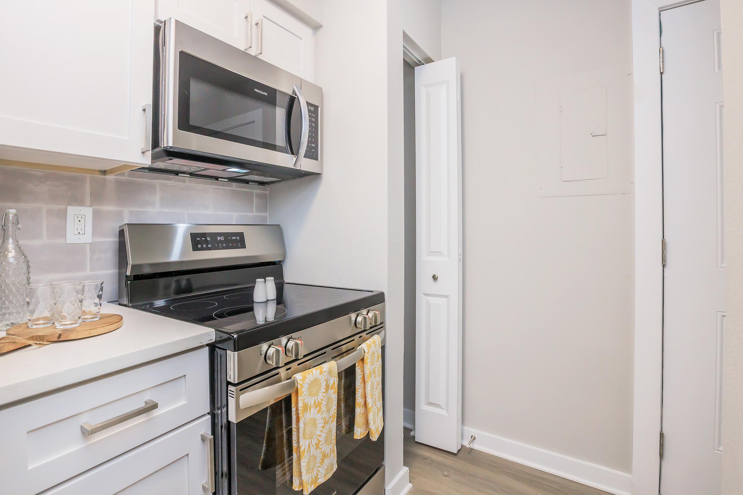 A modern kitchen featuring a stainless steel microwave and oven, white cabinets, and light-colored countertops. A pair of yellow dish towels hang from the oven door. A small table with glassware is on the counter, and there is a door leading to a hallway on the right side. The overall decor is bright and clean.