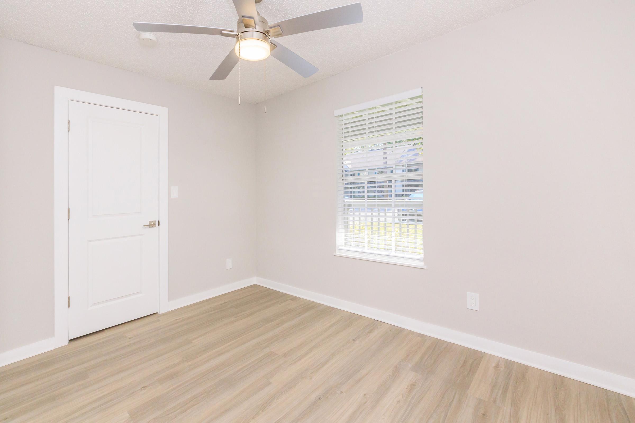 A neutral-colored room with light wood flooring and a ceiling fan. There is a window with blinds letting in natural light, creating a bright atmosphere. A closed door is visible on one side, and the walls are painted in a soft shade, contributing to a minimalist and airy feel.