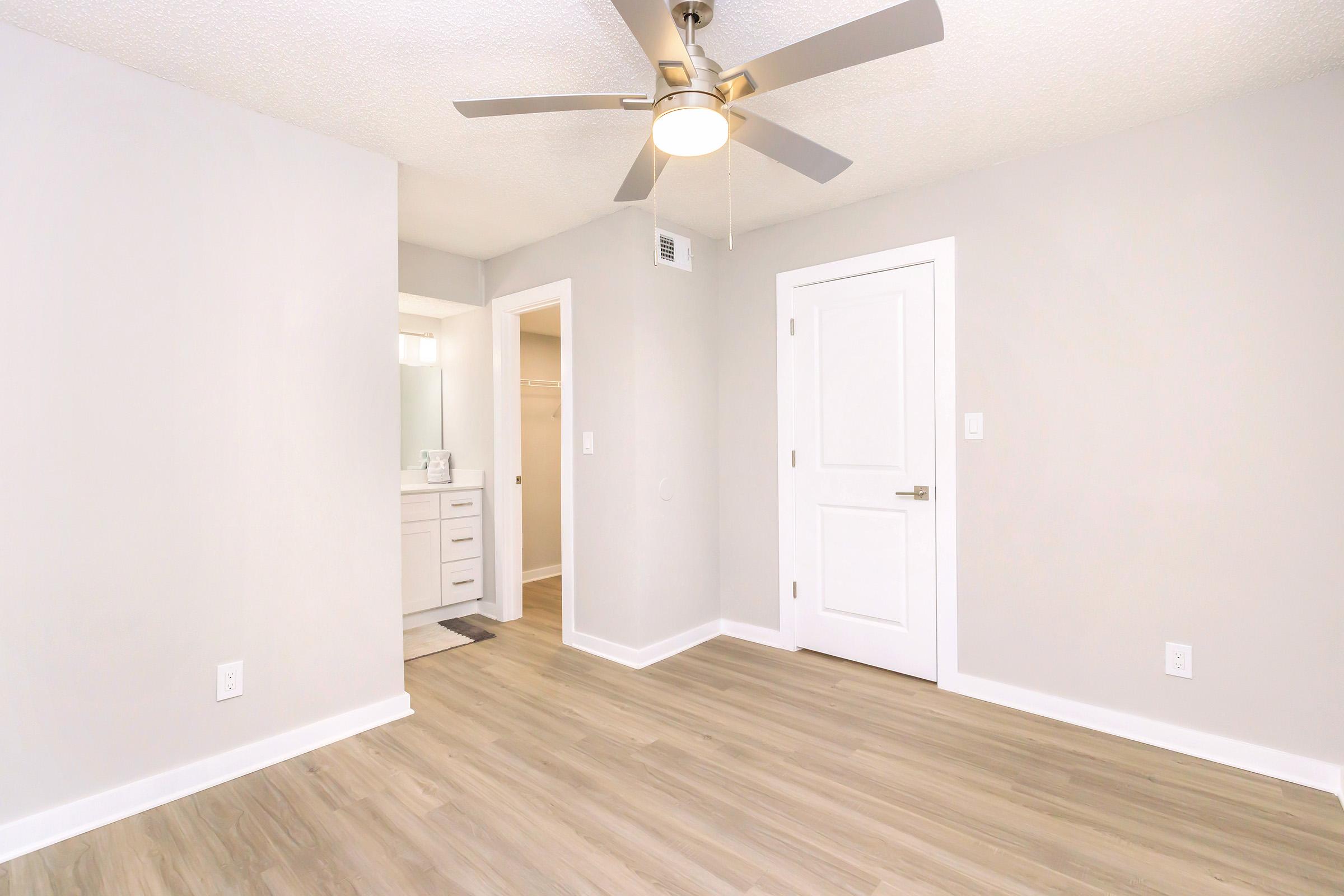 A modern, empty bedroom featuring light gray walls, a ceiling fan, and wood-like flooring. A doorway leads to a bathroom area with a vanity, and there is a white door on the right. The space is well-lit, creating a bright and airy atmosphere.