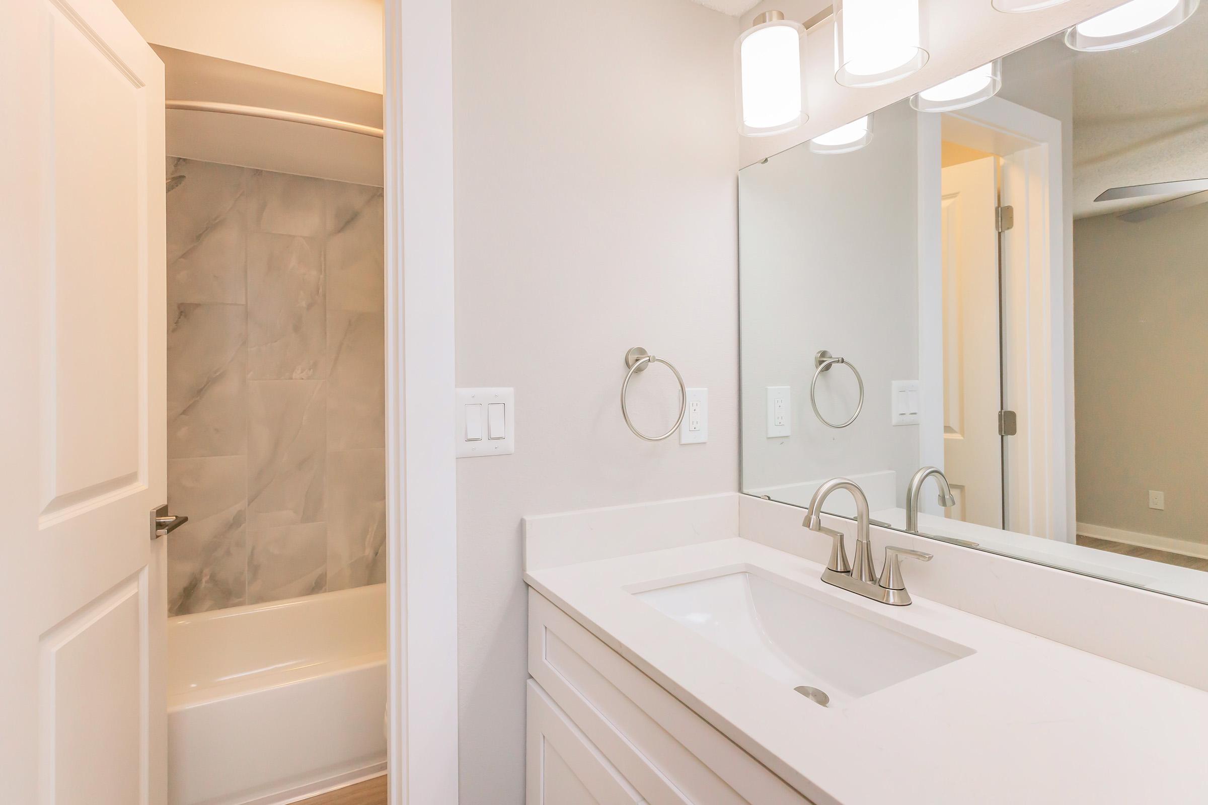 A modern bathroom featuring a light-colored interior with a white vanity and sink. A large mirror is mounted above the sink, reflecting soft lighting from three ceiling fixtures. In the background, a white tub and shower combination is visible, along with a door leading to another area.
