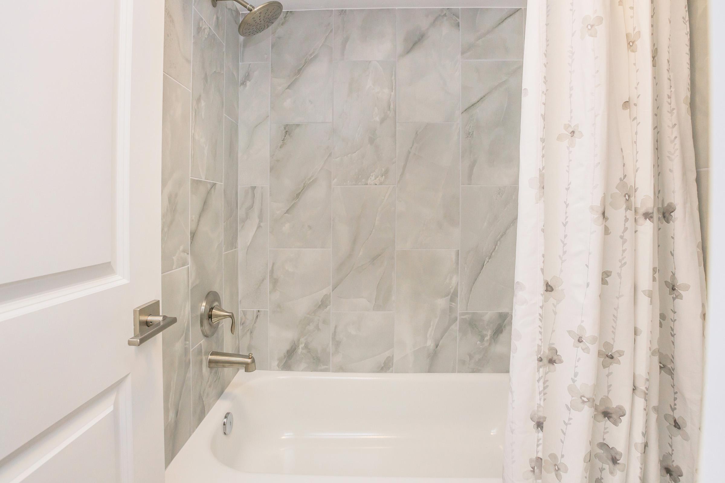 A clean and modern bathroom shower area featuring gray marble tile walls, a chrome showerhead, and a white bathtub. The space is complemented by a light-colored shower curtain with a floral design. A white door is visible on the left side of the image.