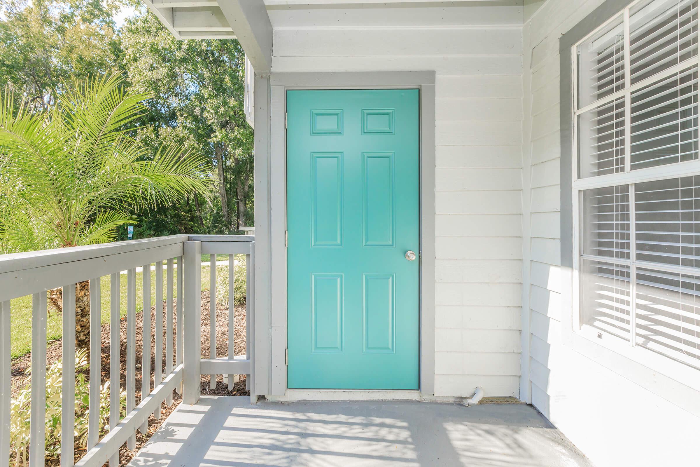 A bright turquoise door with six panels stands on a light-colored house exterior, framed by gray trim. The door is located on a porch with a railing, surrounded by greenery and sunlight filtering through trees. The area features a neat, inviting landscape.