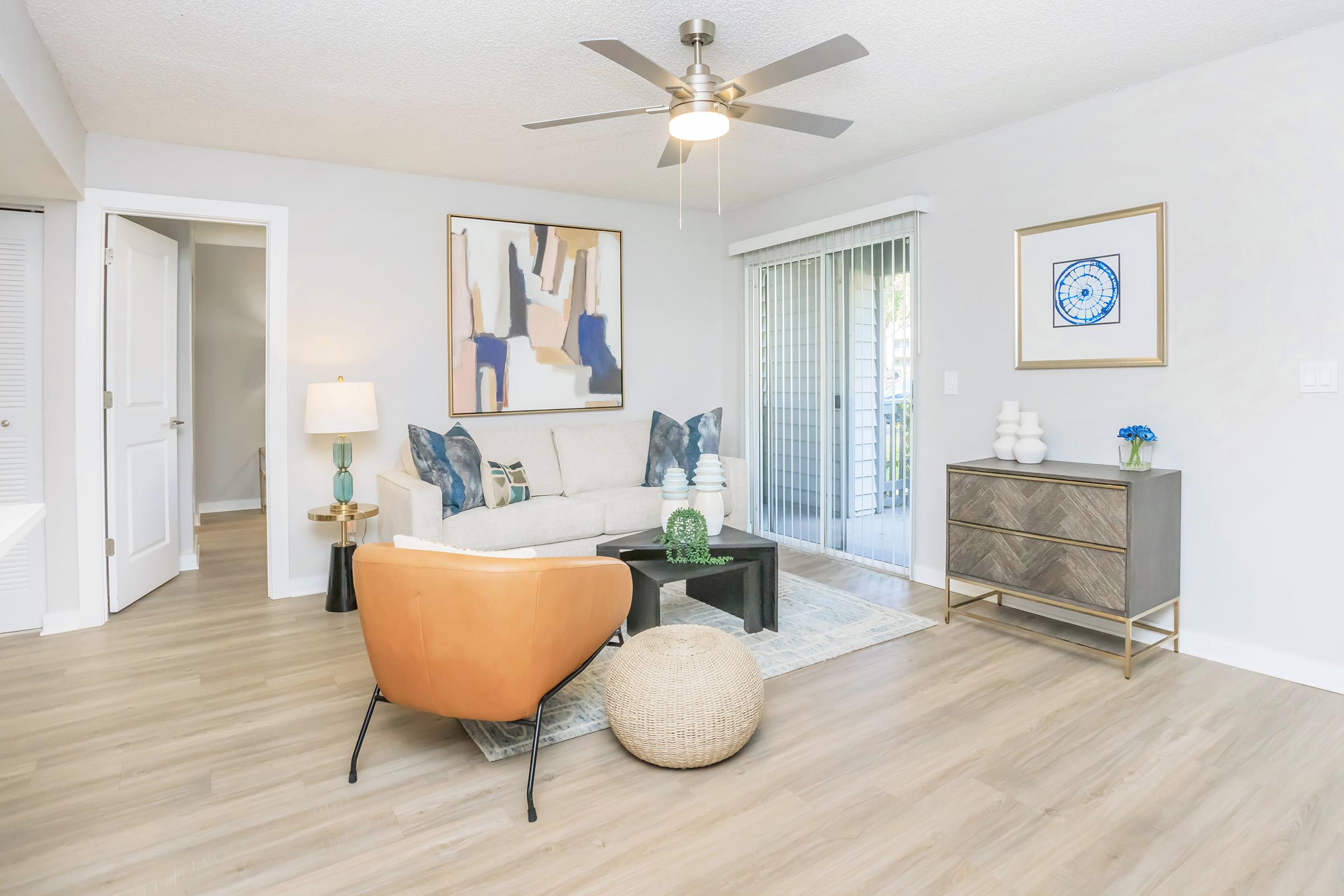 A modern living room featuring a light gray wall, a beige sofa with decorative pillows, an orange armchair, a coffee table with a green vase, and a round pouf. There's a ceiling fan, a sliding glass door with sheer curtains, and artwork on the wall. A stylish cabinet is positioned nearby.