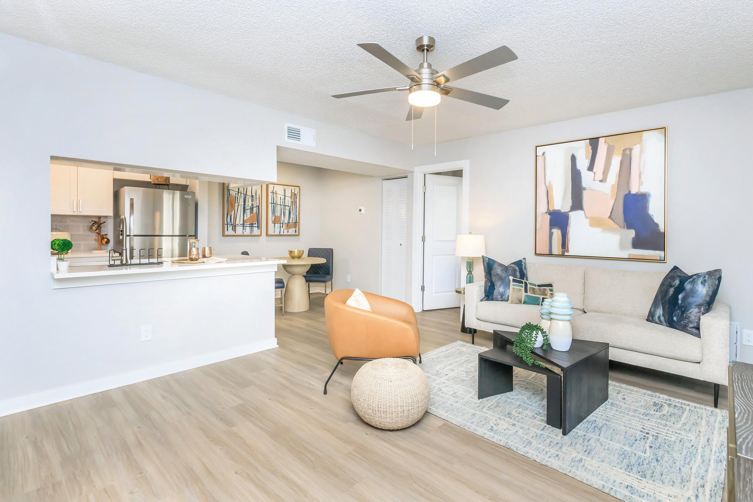 A cozy living room featuring a light-colored sofa with decorative pillows, a wooden coffee table, and a round pouf. In the background, there is a modern kitchen with stainless steel appliances and artwork on the walls. The room is well-lit with a ceiling fan, showcasing a neutral color palette.