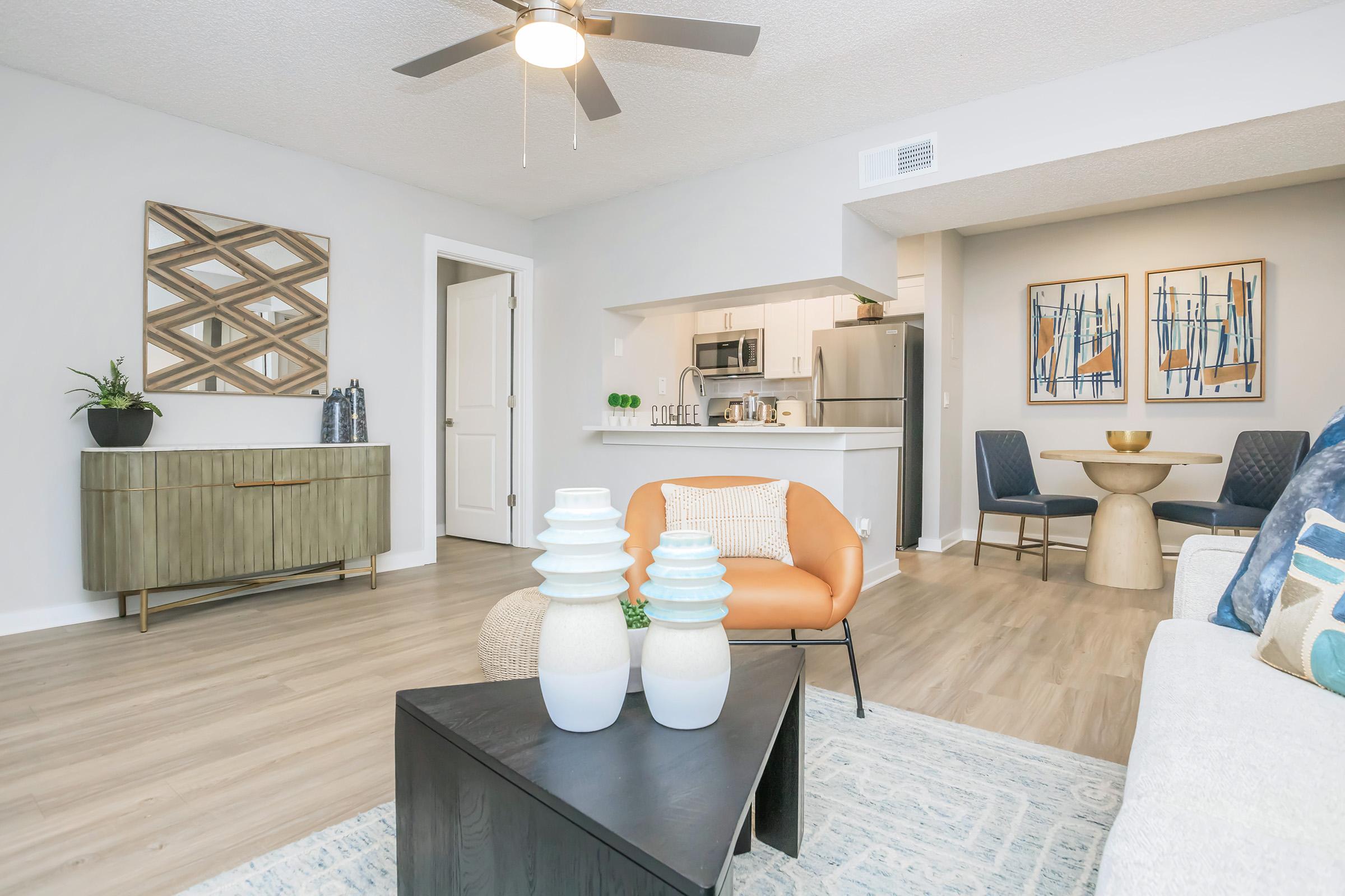 A modern living room featuring a light-colored sofa, an orange accent chair, and a dark coffee table with decorative items. A round dining table with chairs and a kitchen in the background. The space has a fan overhead and decorative wall art, creating a cozy and inviting atmosphere.