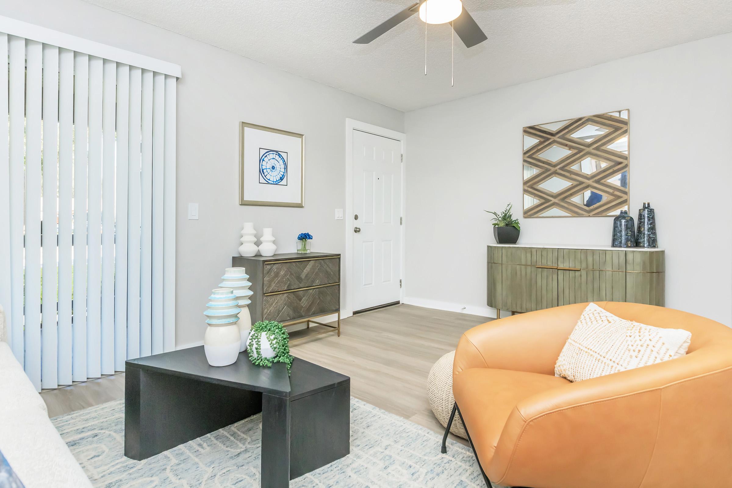 A bright and modern living room featuring a tan leather armchair, a black coffee table, and a decorative rug. The walls are painted light gray, and there's a wall-mounted mirror with geometric designs. Blinds cover a window, and a door leads outside. Decorative plants and vases add a touch of greenery.