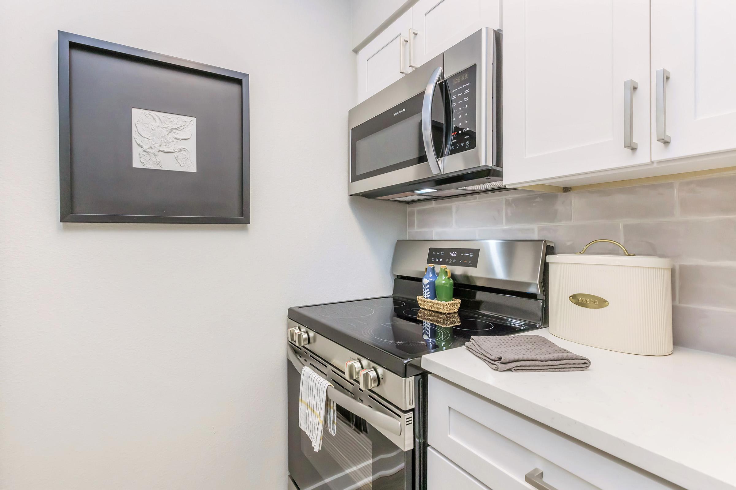 Modern kitchen featuring stainless steel appliances, including a microwave and oven. The cabinetry is white, complemented by a light-colored countertop. A decorative framed artwork is mounted on the wall, and a round, white kitchen canister sits next to a gray dish towel on the counter.