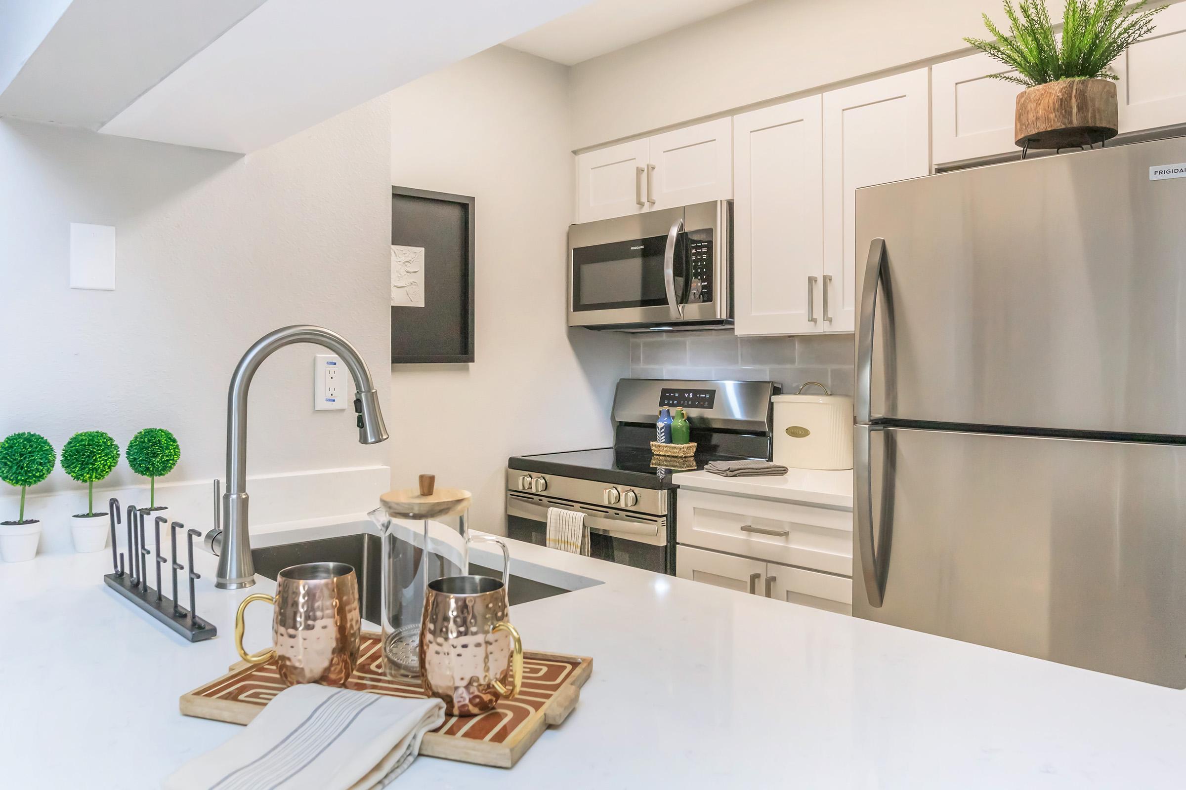 A modern kitchen featuring stainless steel appliances, including a microwave and refrigerator. The countertop is white with a sink and decorative items, including a coffee pot and two copper mugs. Potted greenery adds a touch of nature to the decor. The cabinets are white, creating a bright and clean aesthetic.