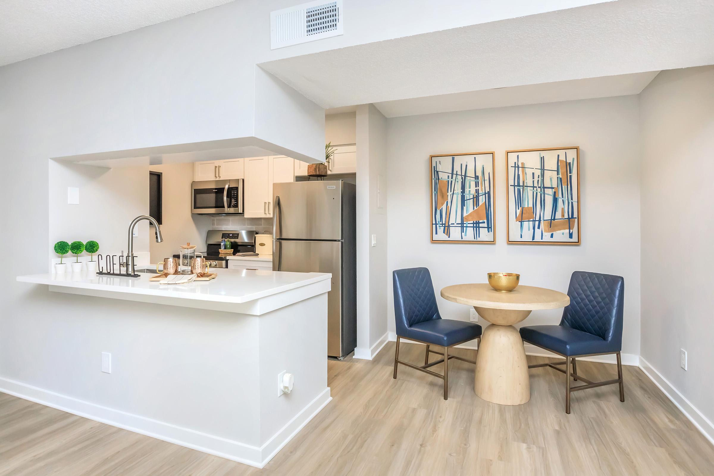 Modern kitchen and dining area featuring a white countertop bar with a sink, stainless steel appliances, and two blue chairs around a round wooden table. Two abstract wall art pieces are displayed, and the floor is light wood. A cozy, inviting atmosphere is created with neutral colors and stylish decor.