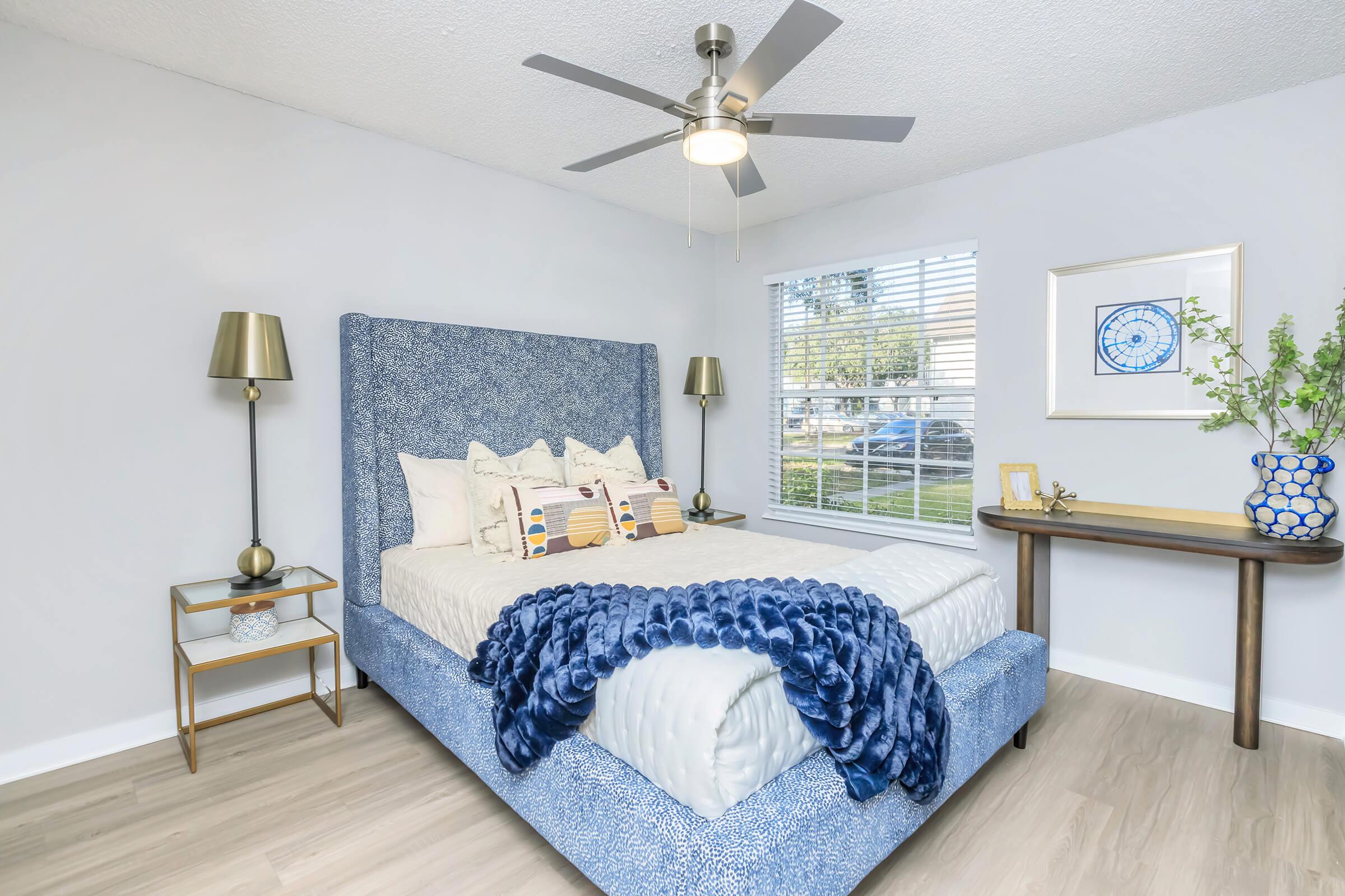 A modern bedroom featuring a blue upholstered bed with decorative pillows, a white and blue throw blanket, two stylish lamps on nightstands, and a small table with decorative items. A window with blinds allows natural light to brighten the space.