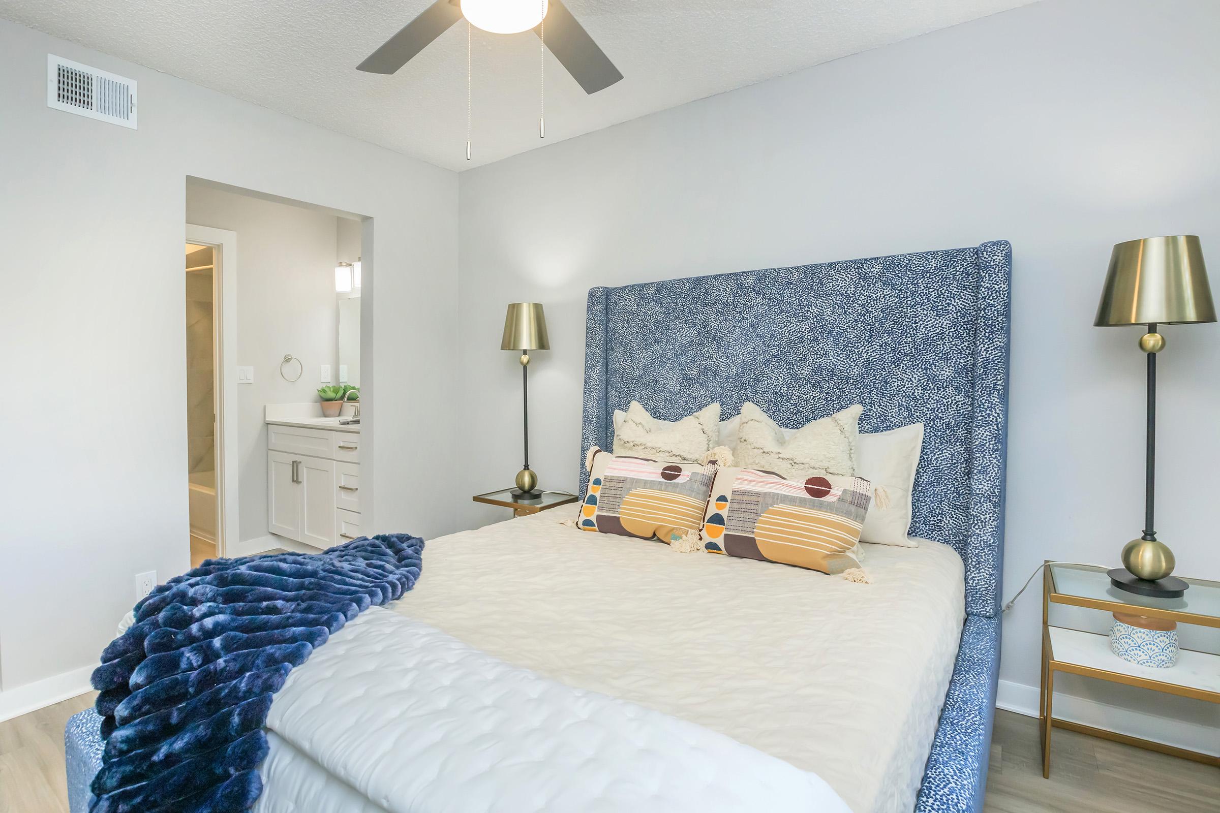 A cozy bedroom featuring a blue patterned headboard with decorative pillows, a white bedspread, and a soft blue blanket. On either side of the bed are modern bedside tables with stylish lamps. A doorway leads to a bathroom, and the wall is painted a light gray, adding to the serene atmosphere.