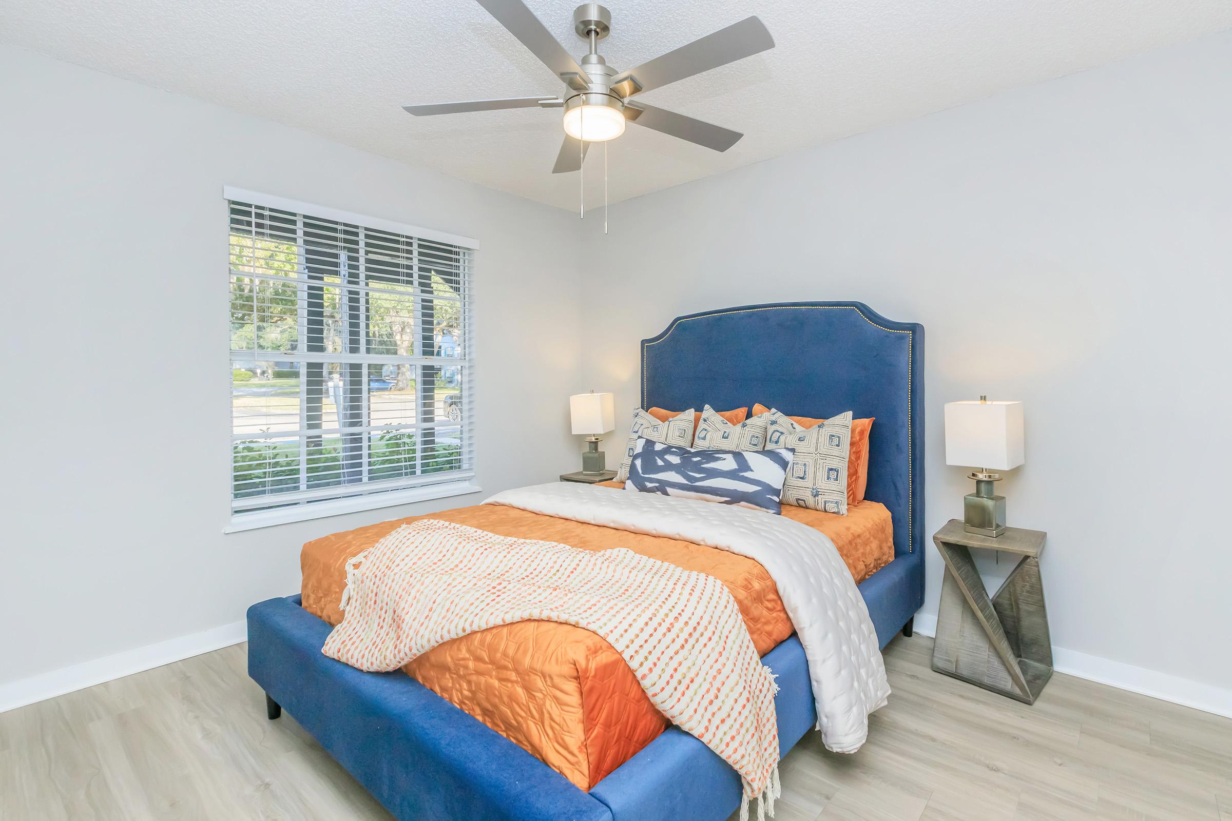 A cozy bedroom featuring a blue upholstered bed with orange and white bedding, decorative pillows, and a stylish nightstand. There are two lamps on either side of the bed, and large windows allow natural light to brighten the space. The walls are painted light gray, and the floor is a light wooden finish.