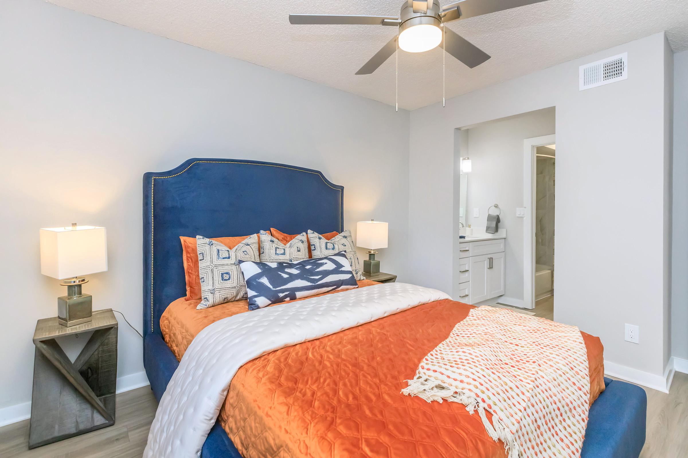 A well-decorated bedroom featuring a blue upholstered bed with patterned pillows and an orange blanket. Two bedside lamps sit on wooden tables, and there is a light-colored wall with a ceiling fan above. A bathroom area is visible in the background, adding to the modern aesthetic.