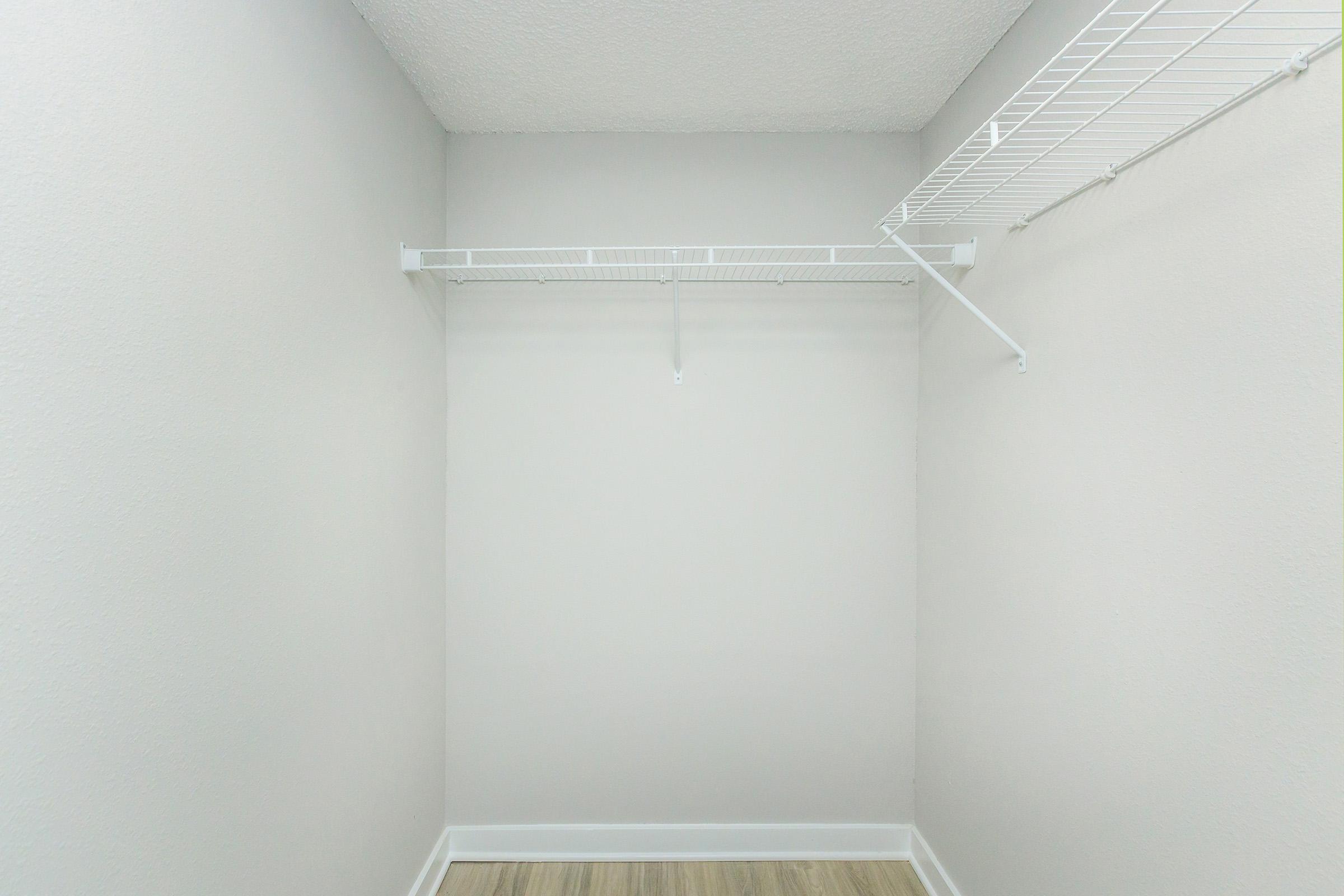 Empty closet with bare walls and a white wire shelving unit mounted on one side, featuring a single hanging rod. The floor is light-colored, creating a bright and airy appearance. The overall ambiance is minimal and uncluttered.