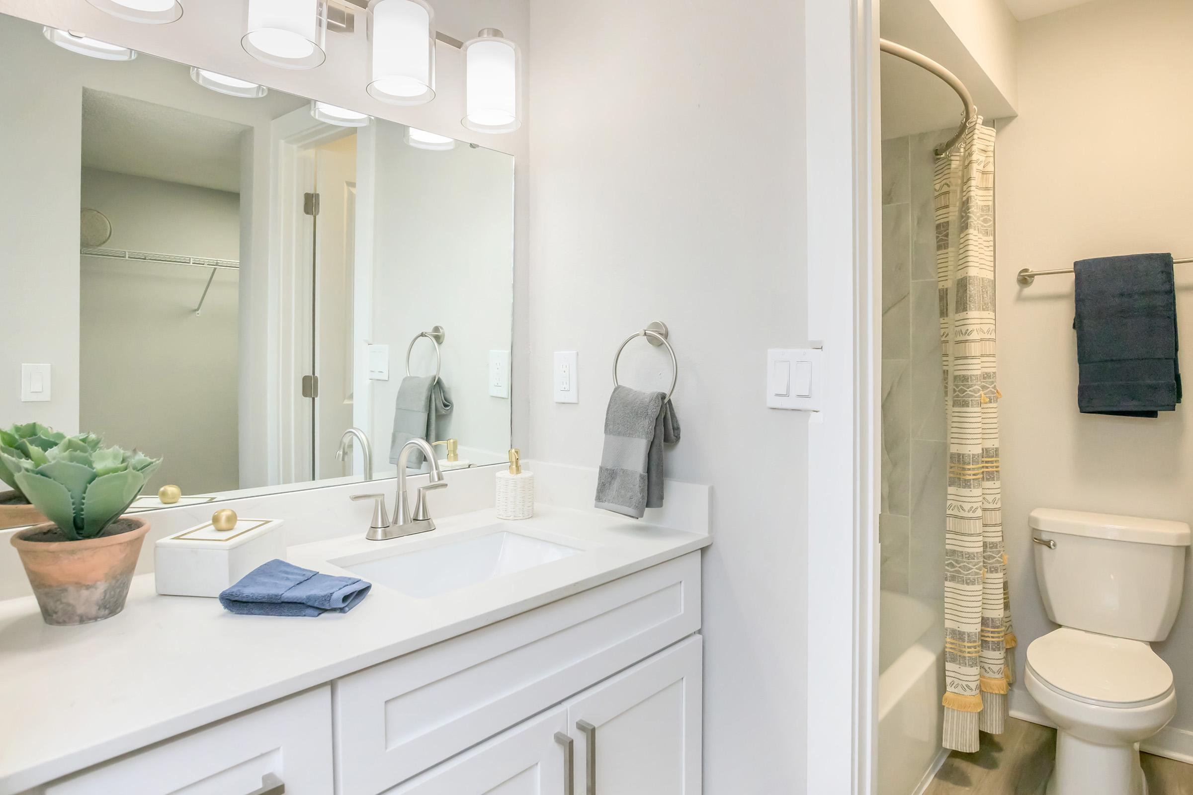 A modern bathroom featuring a white vanity with a sink, stylish light fixtures, and a mirror. There is a shower with a curtain, a toilet, and a potted plant on the countertop. Soft towels hang nearby, creating a clean and inviting appearance.