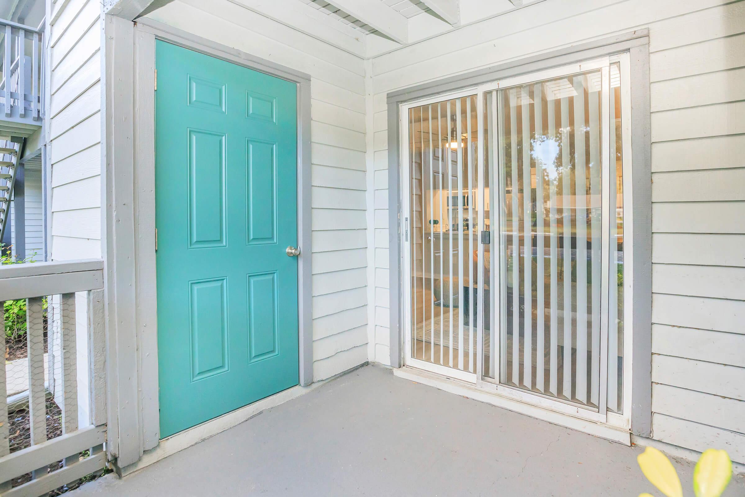 A light-colored exterior featuring a teal door on the left and a sliding glass door with vertical blinds on the right. The entrance is surrounded by white siding and has a concrete floor. Green foliage is visible in the background, suggesting a pleasant outdoor setting.