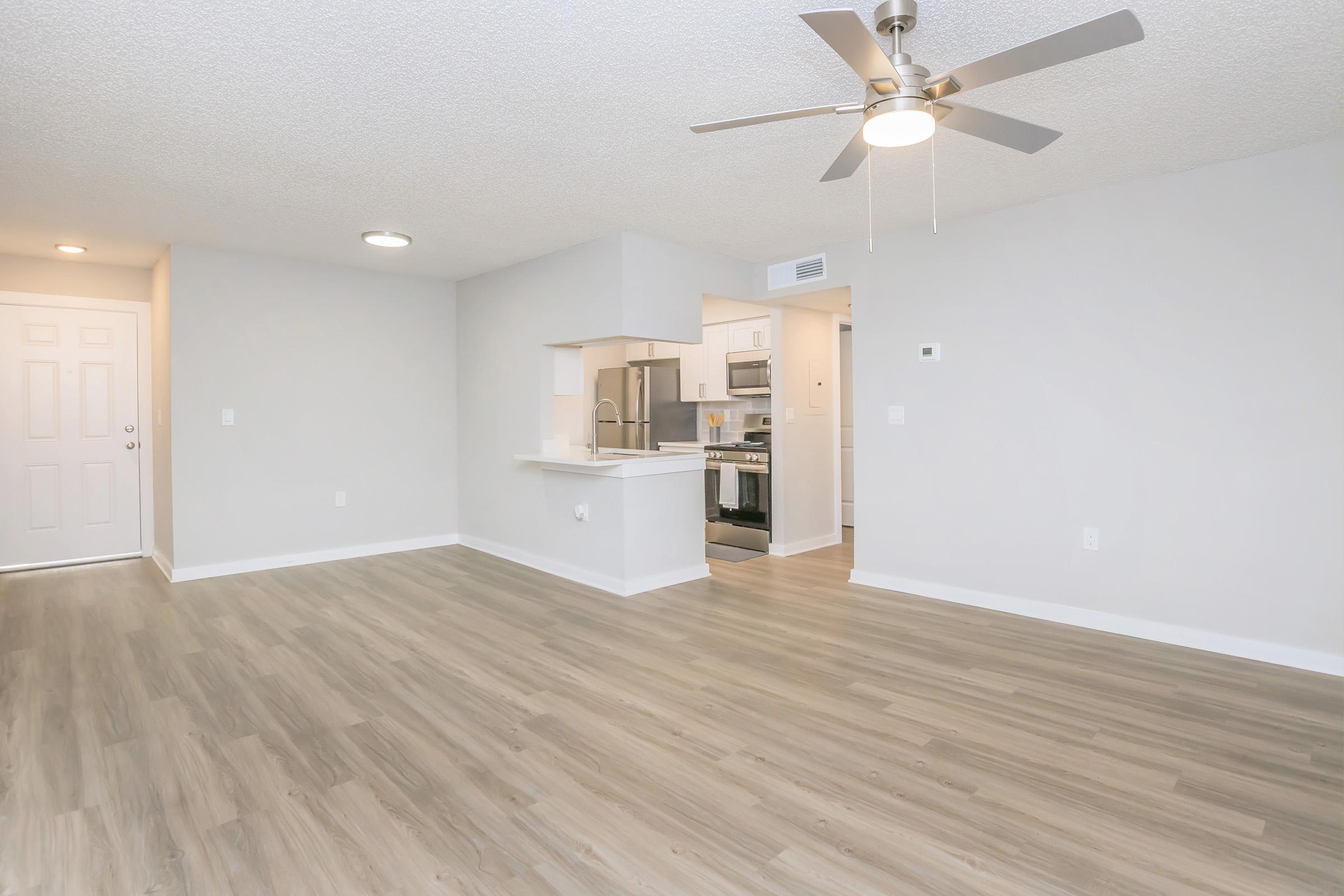 A spacious, well-lit living area with light-colored walls and wooden flooring. A ceiling fan hangs from the ceiling. In the background, a kitchen is visible, featuring modern appliances and a breakfast bar. A doorway leads to another room, while the front door is to the left.
